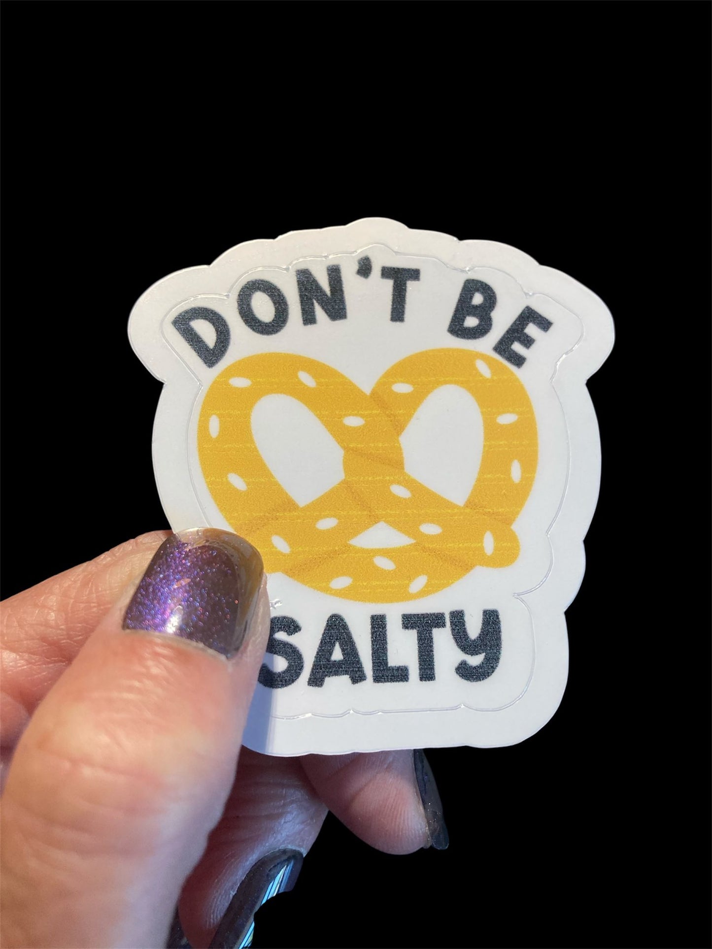 Sticker with pretzel design and text 'Don't Be Salty' held by a hand against a black background