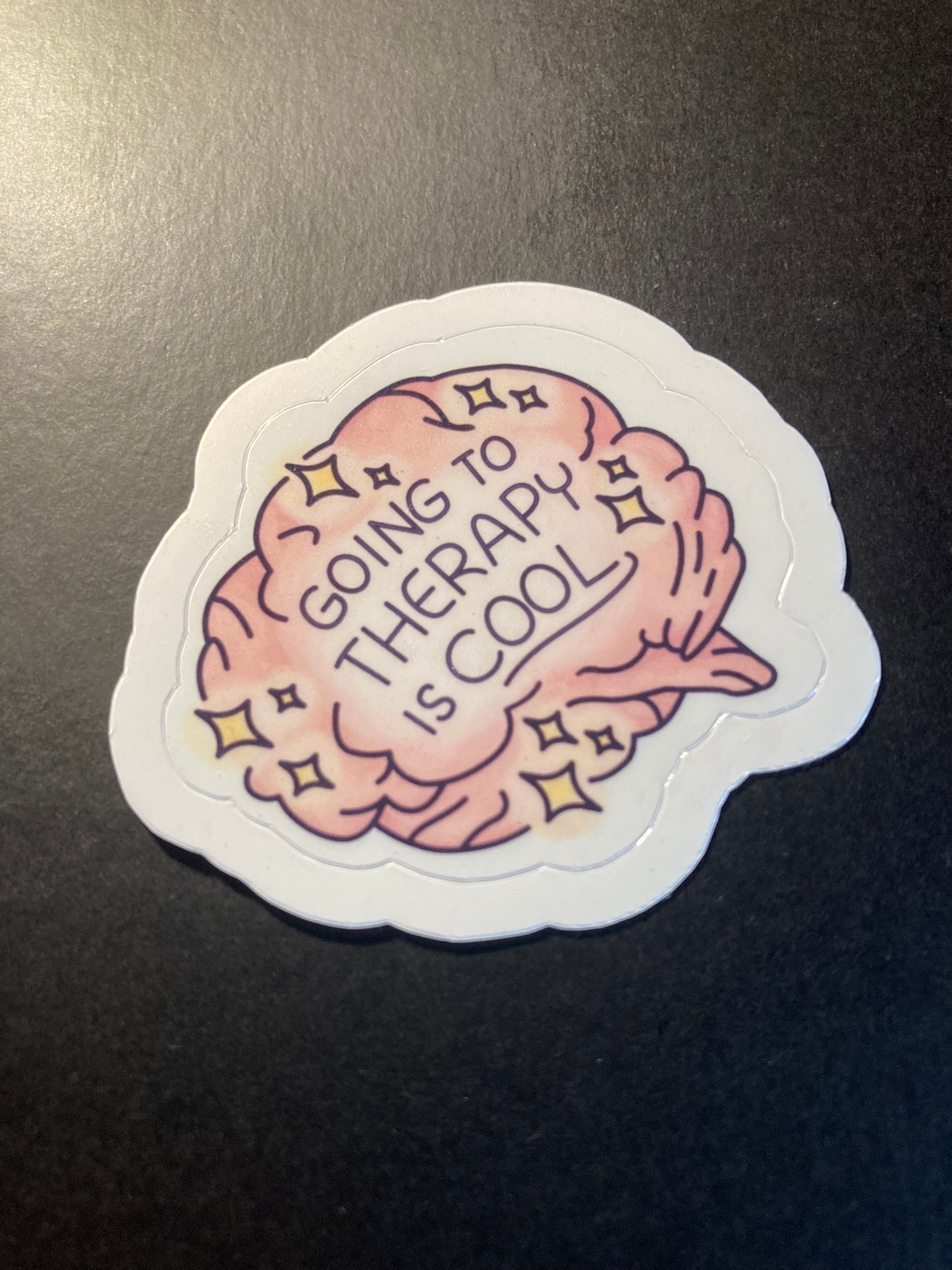 Sticker with brain design and text 'Going to therapy is cool' on a dark background