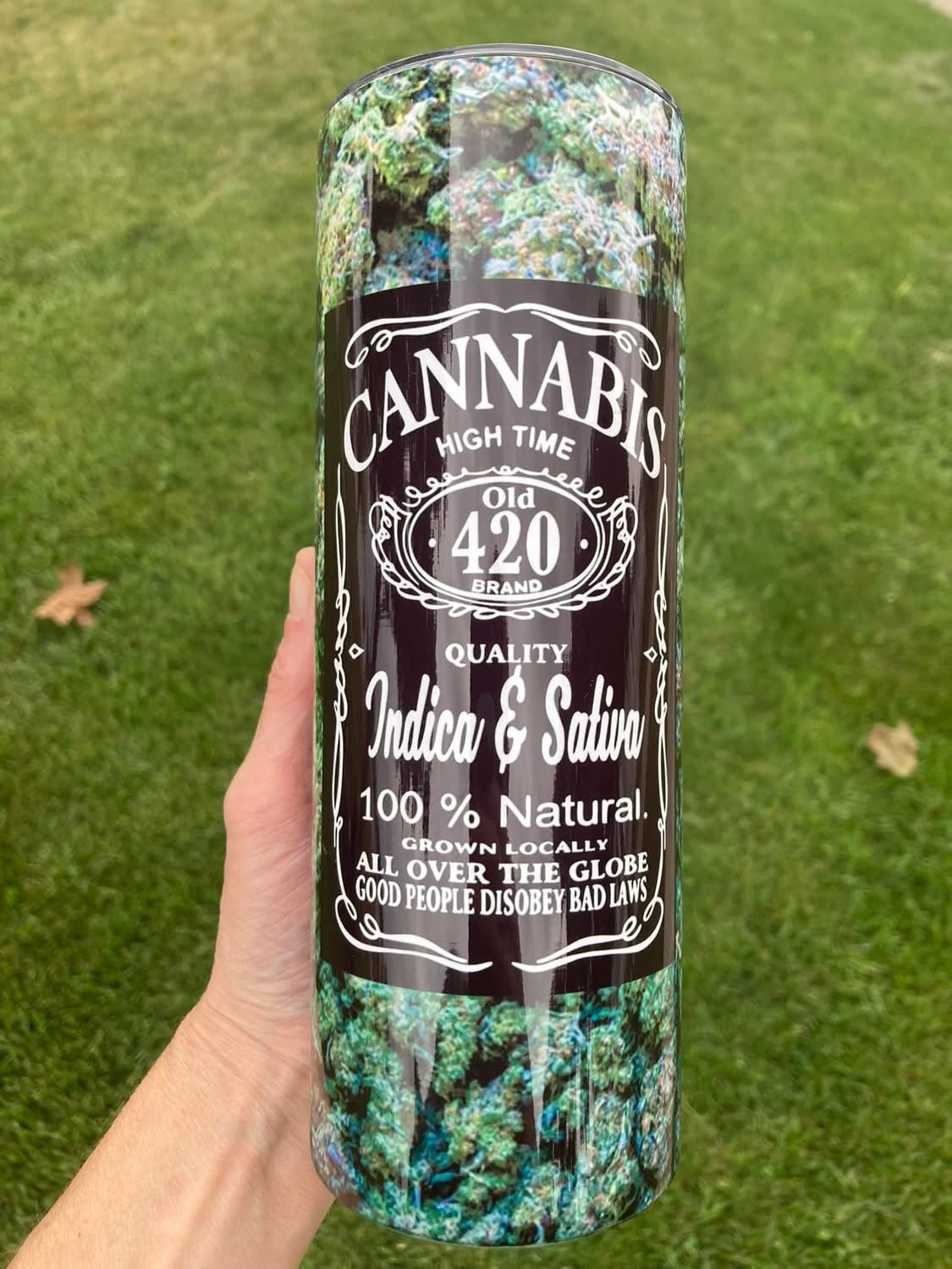 Tumbler with cannabis-themed design held against a grassy background. 20oz skinny tumbler featuring cannabis buds background with vintage 420 label design reading Indica and Sativa, held outdoors on grass