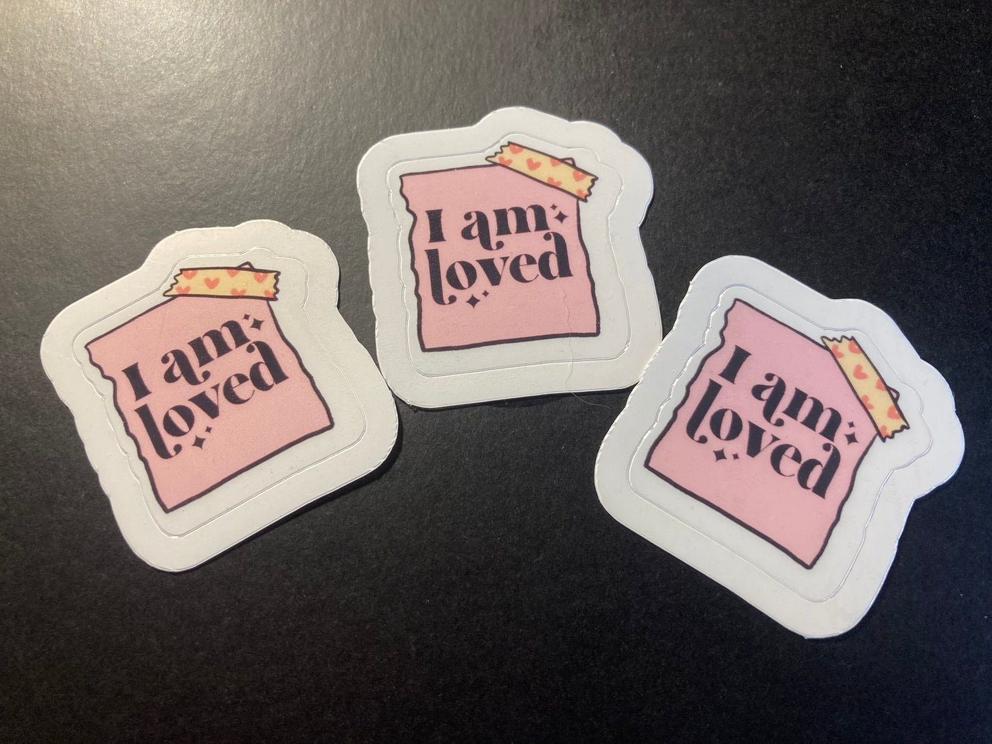 Three 'I am loved' stickers on a dark surface