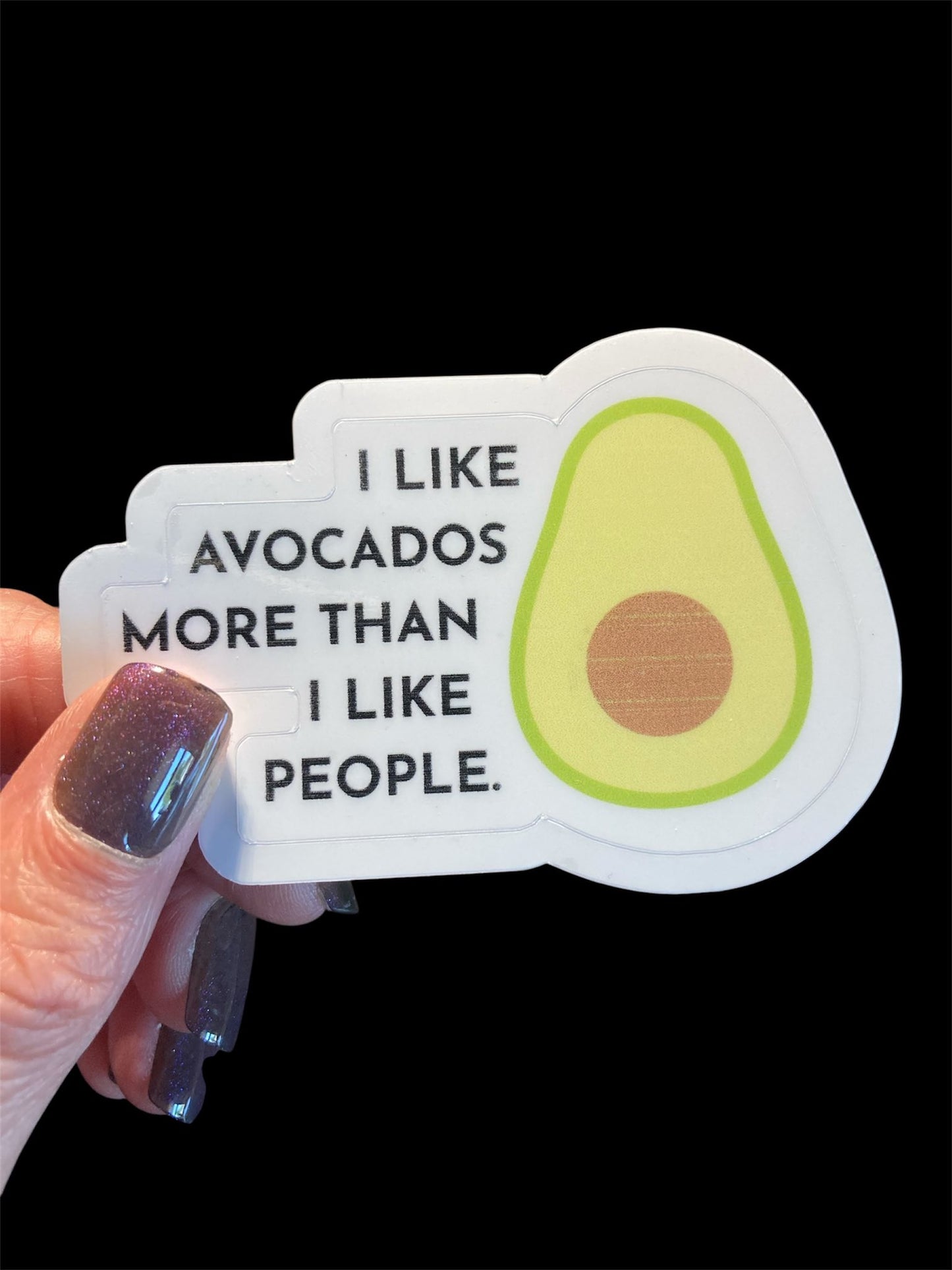 Sticker with avocado graphic and text held by a hand against a black background