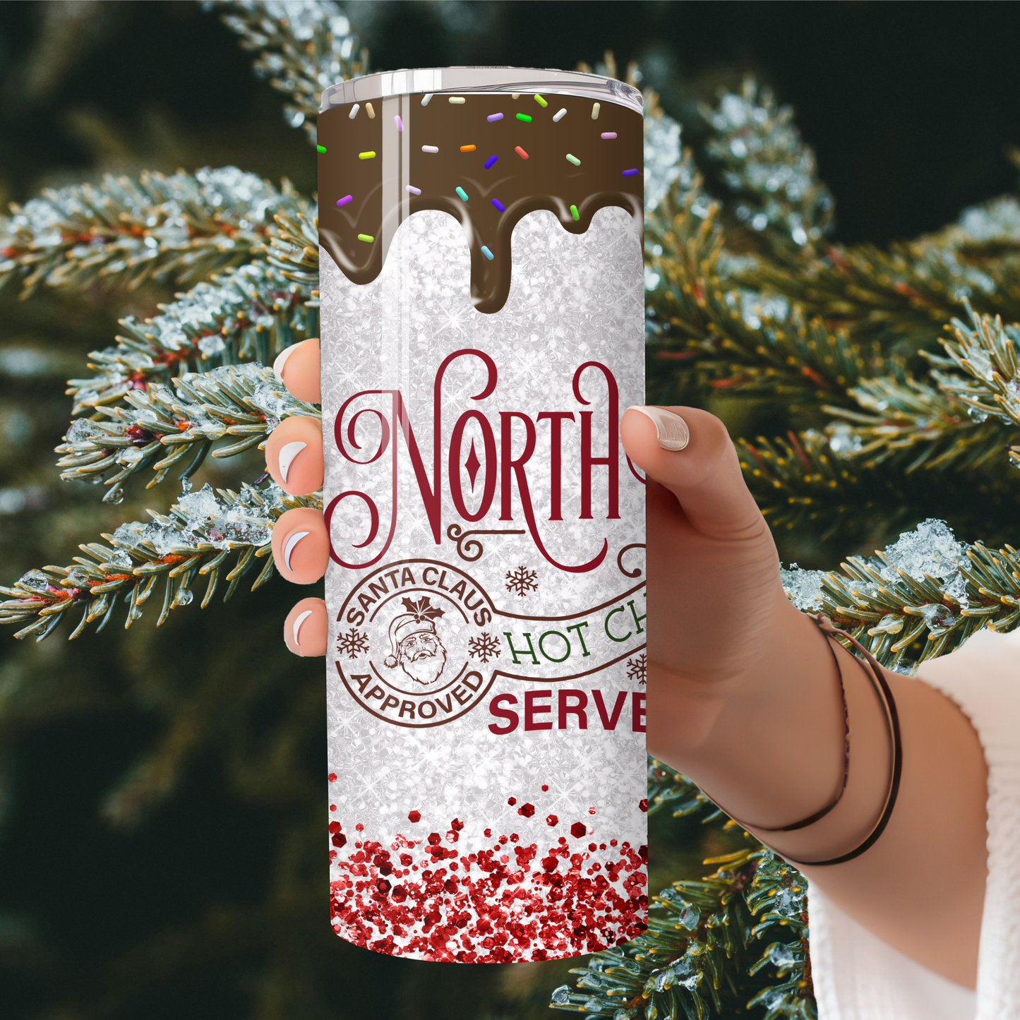 Tumbler with 'North Pole Hot Chocolate' design held in front of a Christmas tree.