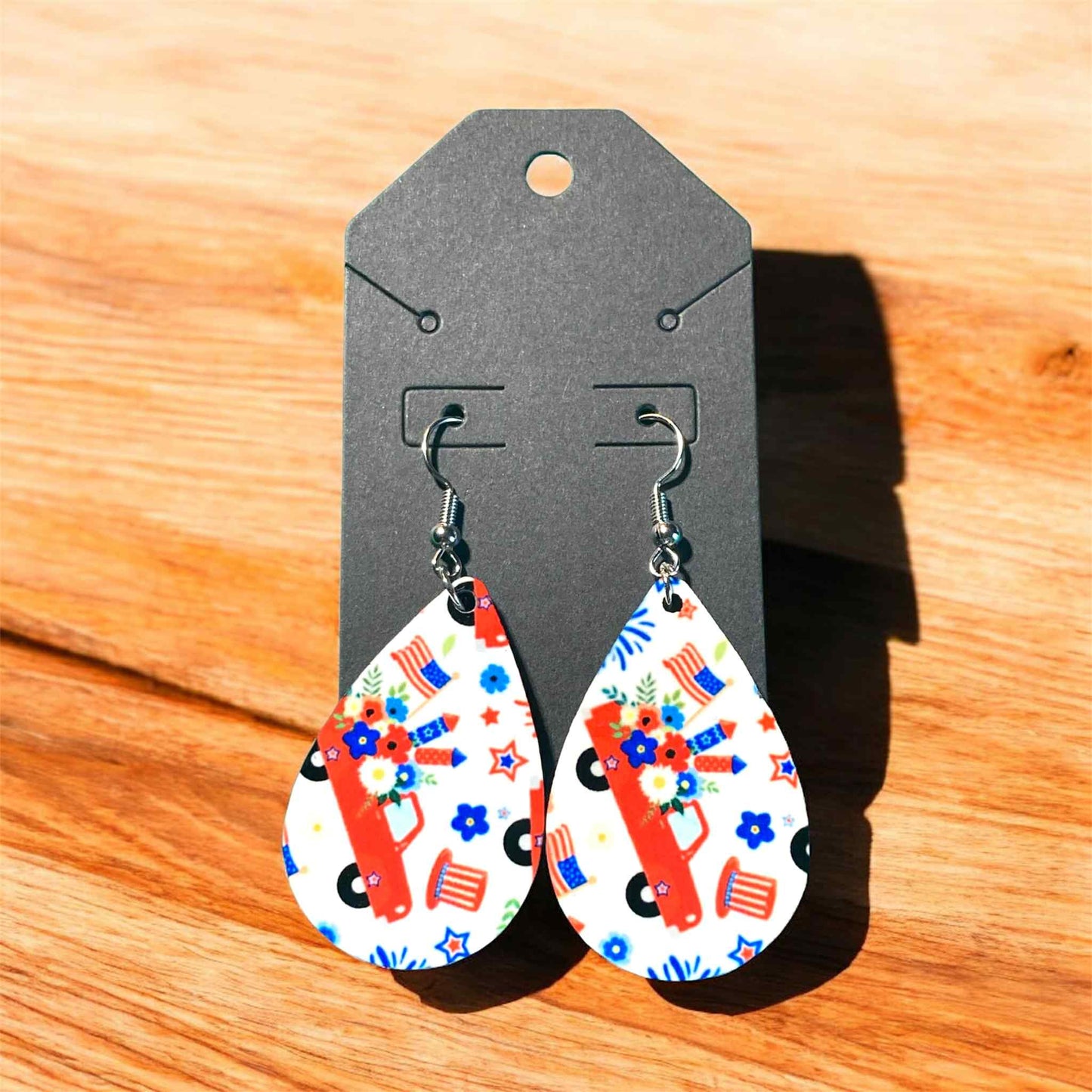 Teardrop-shaped earrings with floral and car design on a gray card against a wooden background. Patriotic Red Truck Floral Teardrop Earrings – Americana Gift