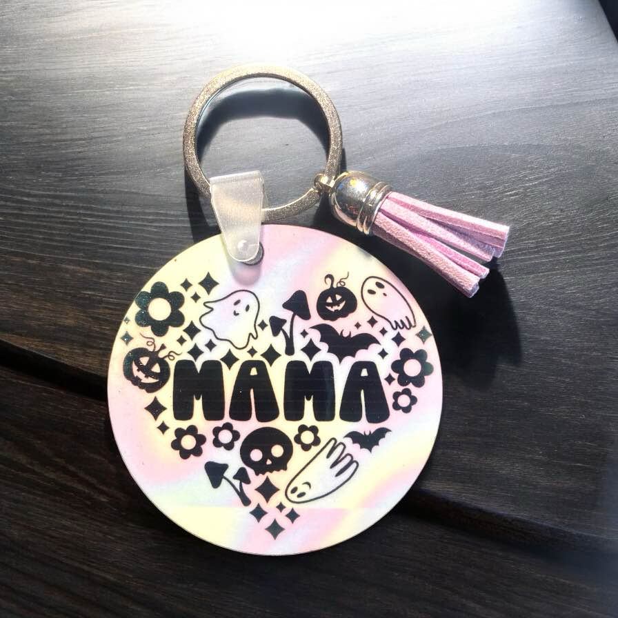 2.5 x 2.5 in circle sublimation keychain. Iridescent hologram background. Image is black. Design phrase says Mama and the images surrounding are in a heart and are ghosts, pumpkins, and checker board. Tassel color is chosen at random.