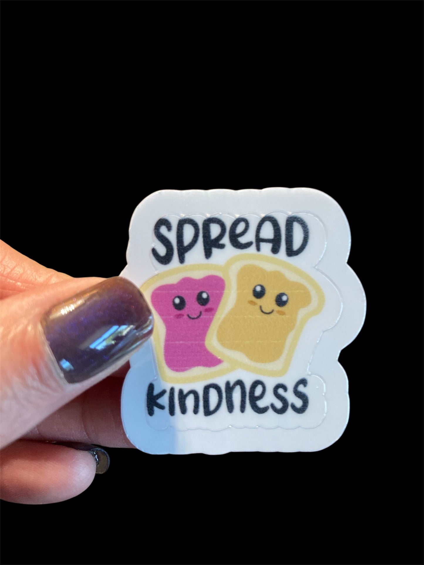 Sticker with cartoon characters and 'Spread Kindness' text held by a hand against a black background