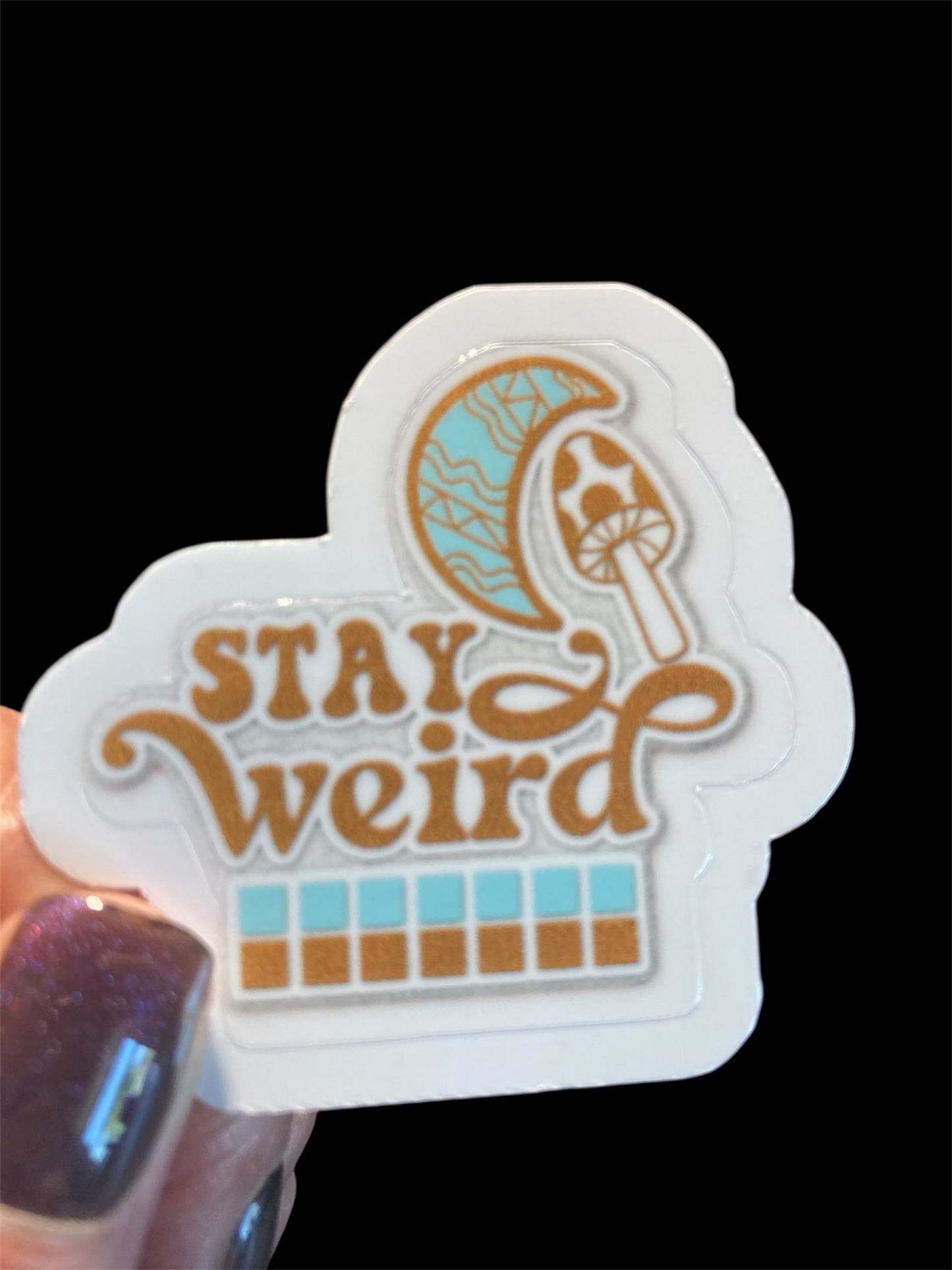 Sticker with 'Stay Weird' text and mushroom design held by a hand.