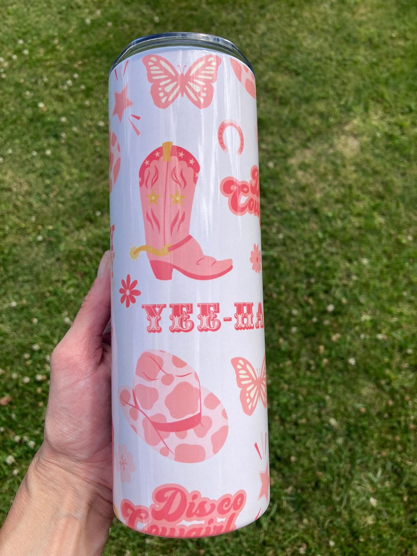 Tumbler with pink cowboy boot and butterfly design held in a hand against a grass background
