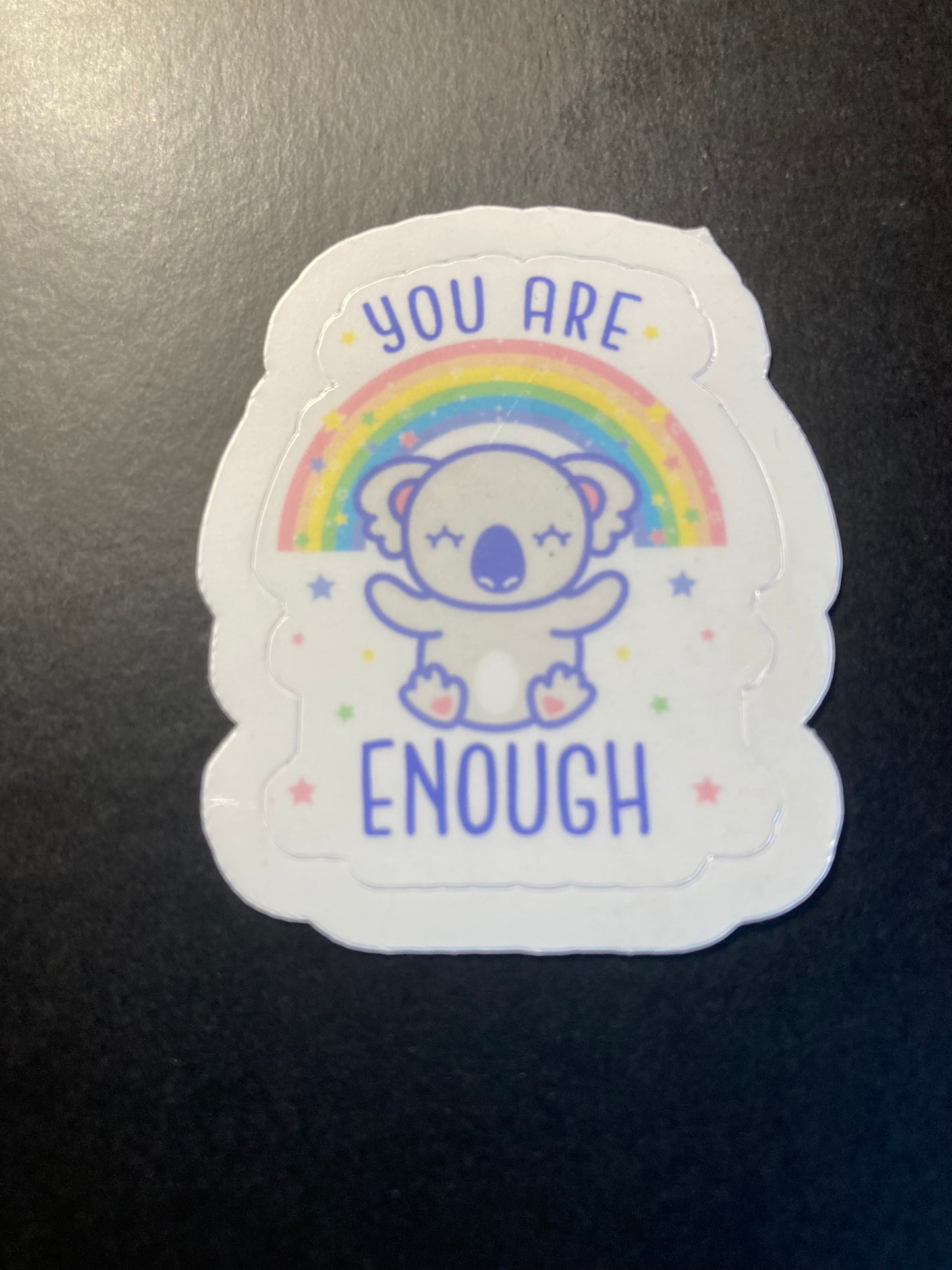 Sticker with a bear illustration, rainbow, and 'You are enough' text on a black surface.