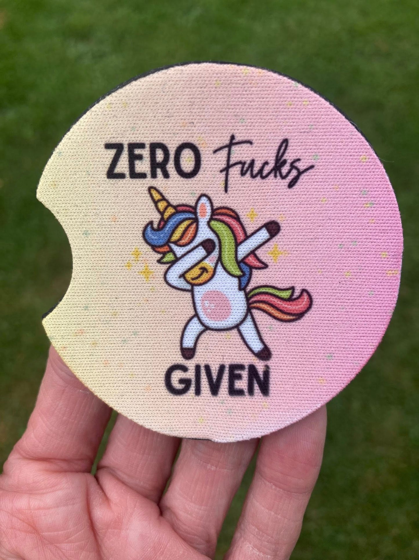 Circular product with unicorn illustration and text 'ZERO Fucks GIVEN' held in a hand against a grassy background.