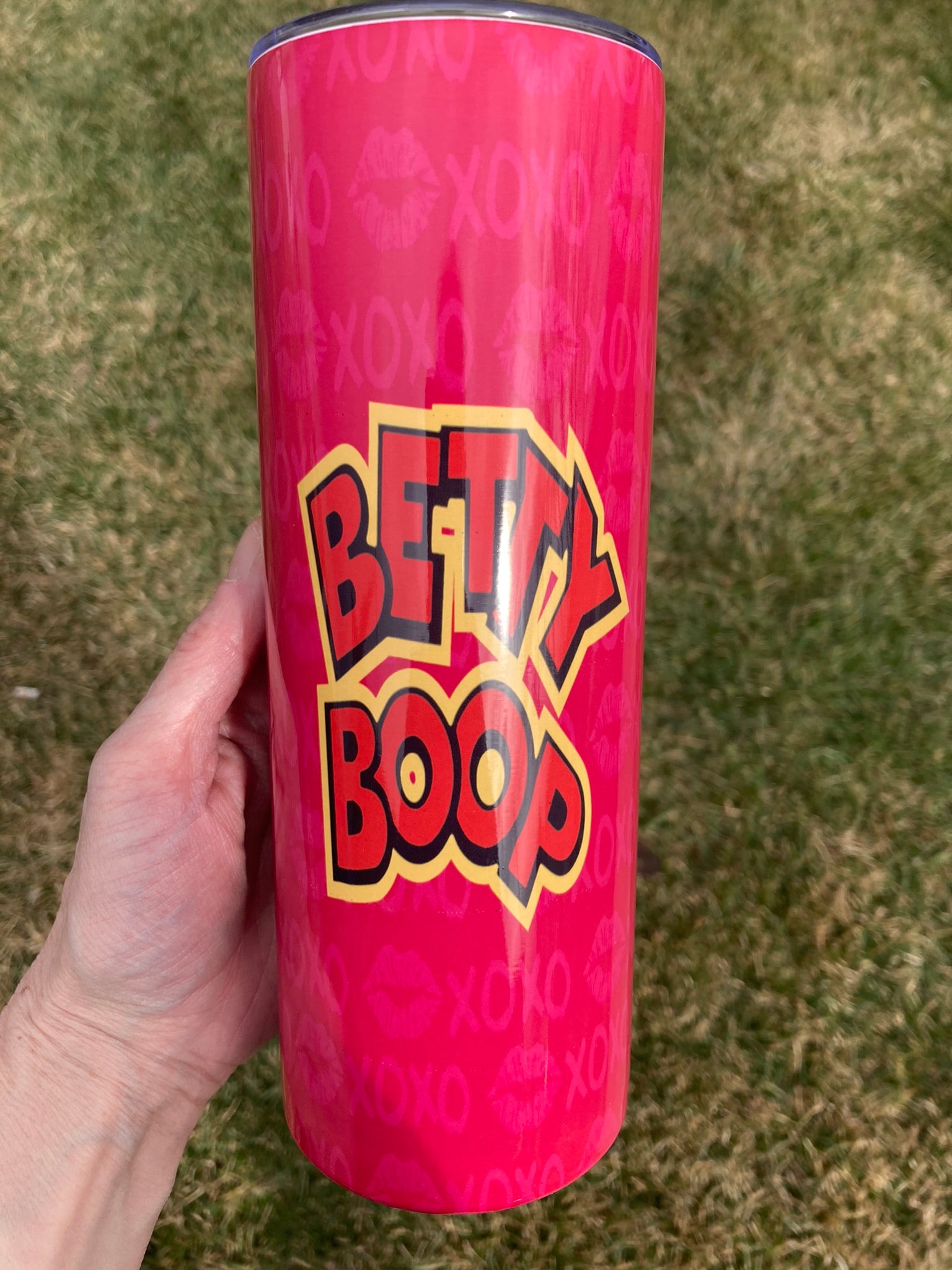 Pink tumbler with 'Betty Boop' branding held by a hand on a grassy background