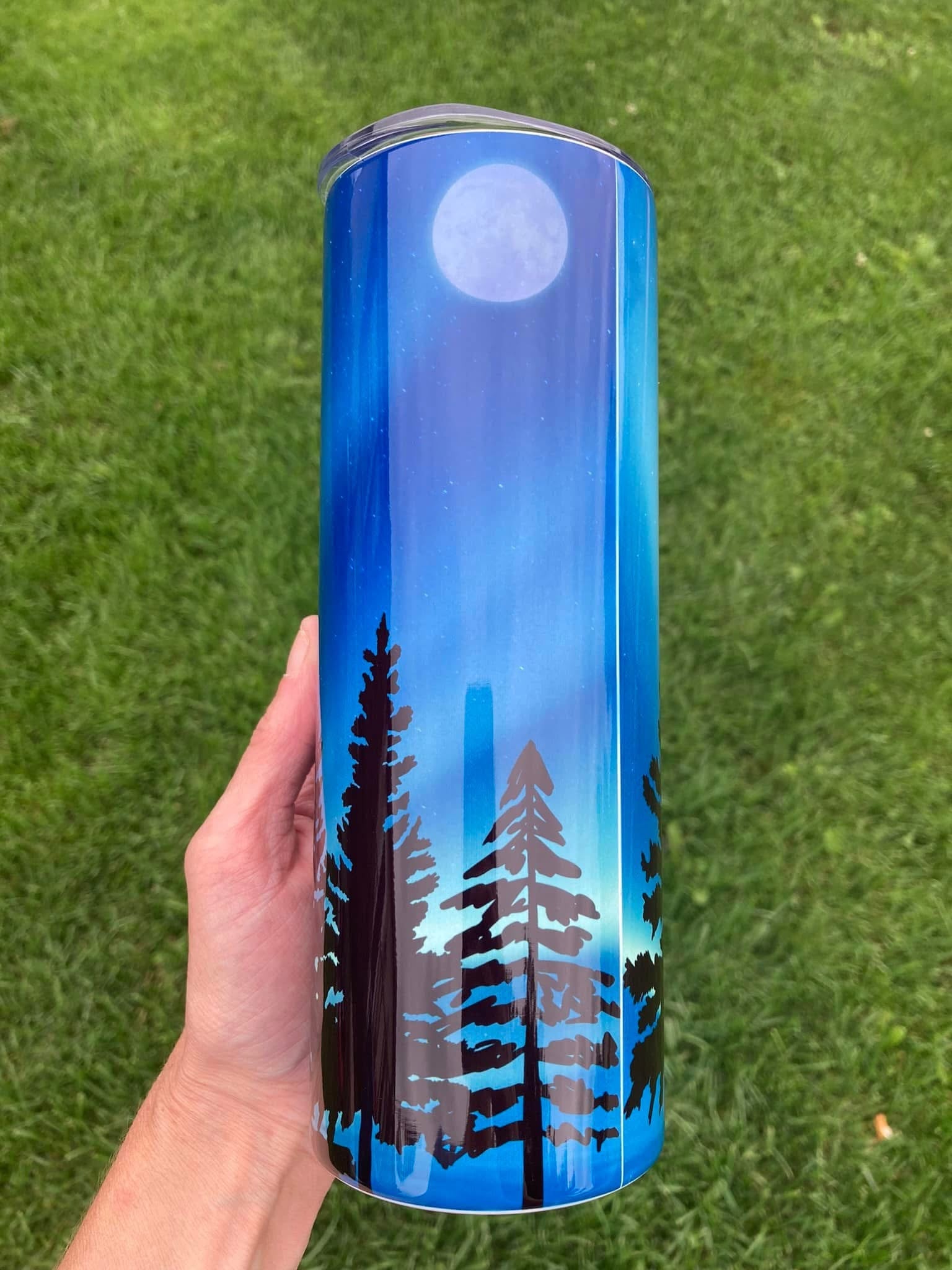 Tumbler with nature design held in hand on grass. 20oz skinny tumbler with blue forest background featuring silhouette of Bigfoot walking among trees