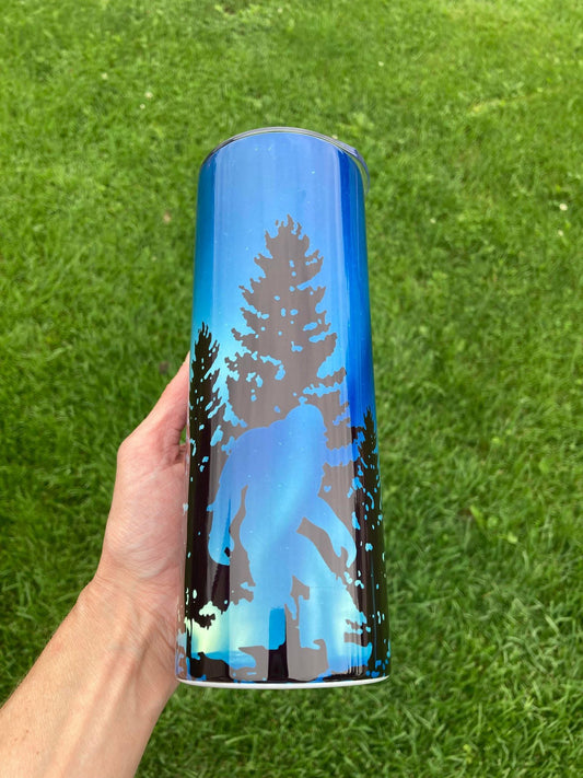 Tumbler with nature design held in front of green grass. 20oz skinny tumbler with blue forest background featuring silhouette of Bigfoot walking among trees