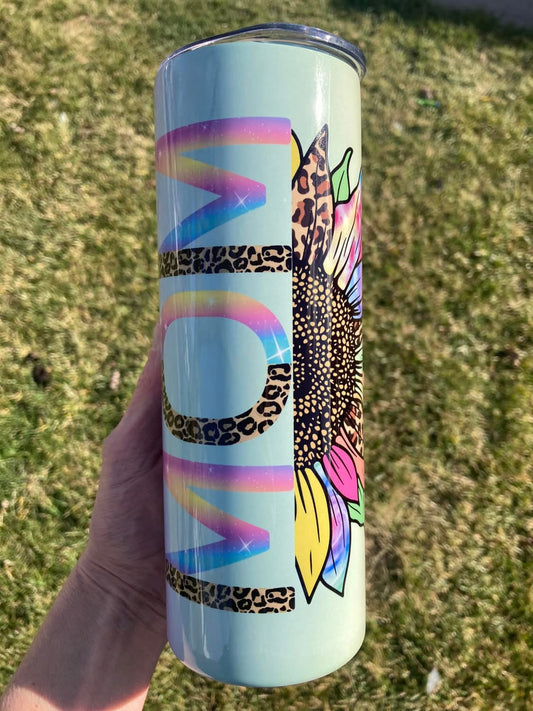Tumbler with colorful design held in a hand against a grassy background. 20oz skinny tumbler with pastel background featuring rainbow letters spelling MOM with leopard print accents and sunflower design