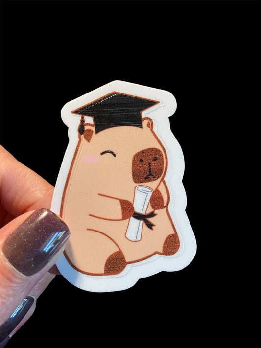 Sticker of a bear in a graduation cap and gown holding a diploma, held by a hand with glittery nail polish on a black background.