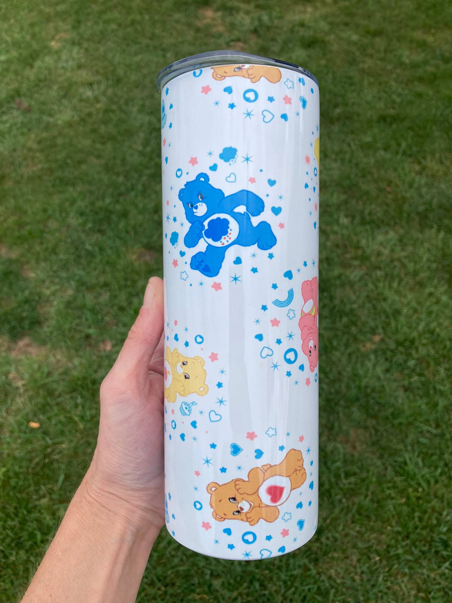 Tumbler with cartoon bear designs held by a hand on a grassy background 20oz skinny tumbler with colorful bear characters and rainbow accents in a cute cartoon aesthetic pattern
