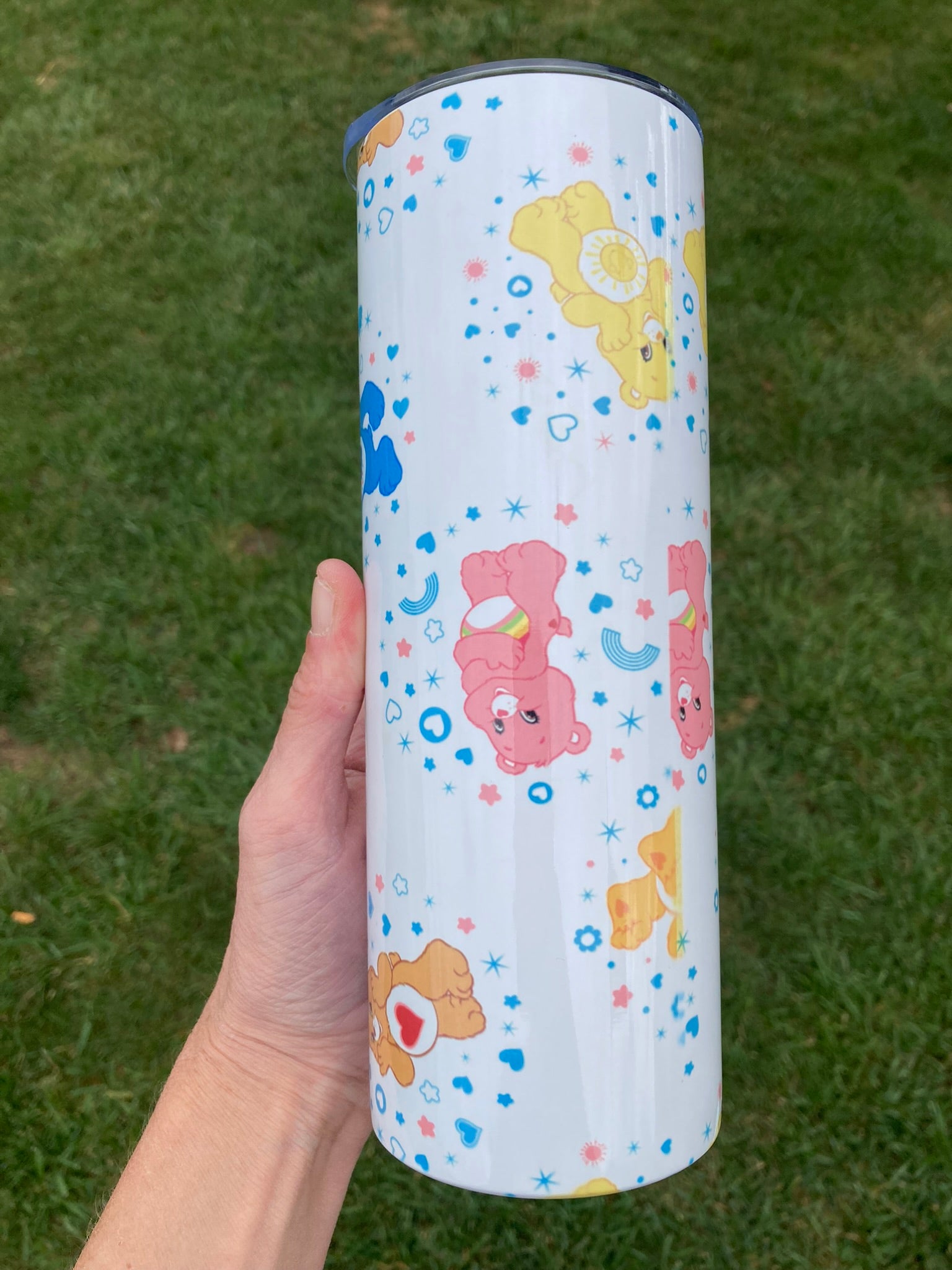 Tumbler with cartoon bear designs held by a hand on a grass background. 20oz skinny tumbler with colorful bear characters and rainbow accents in a cute cartoon aesthetic pattern