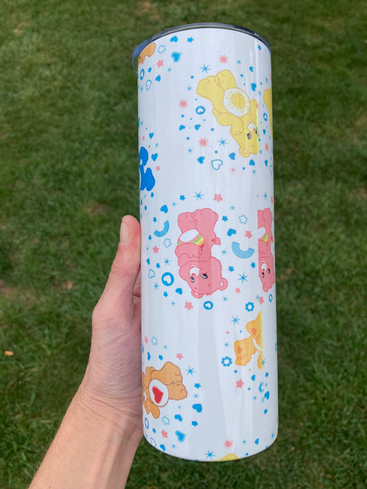 Tumbler with cartoon bear designs held by a hand on a grass background. 20oz skinny tumbler with colorful bear characters and rainbow accents in a cute cartoon aesthetic pattern