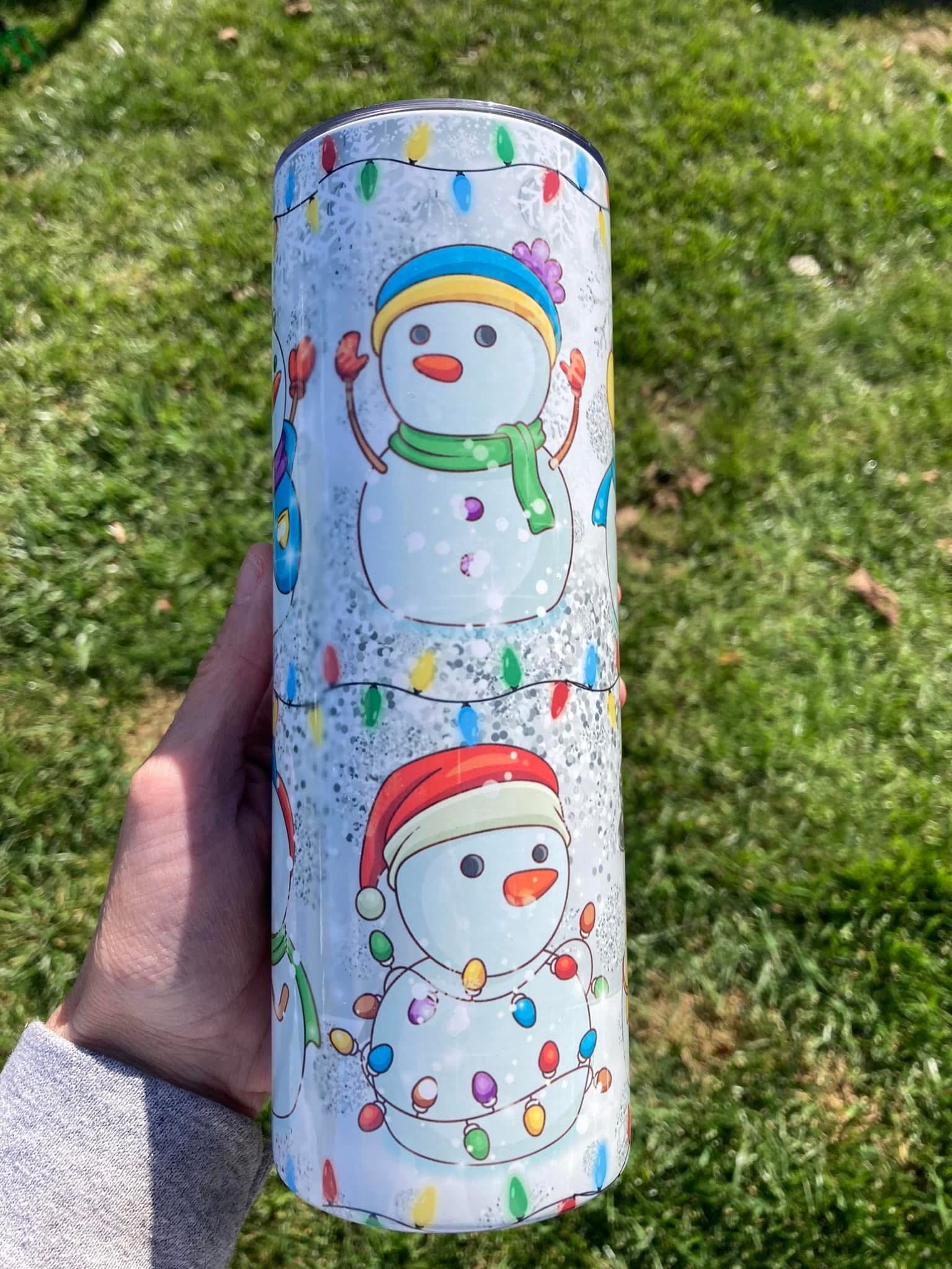 Tumbler with snowman design held in front of grass. Snowman Holiday Christmas Tumbler – Holiday Lights Winter Drinkware