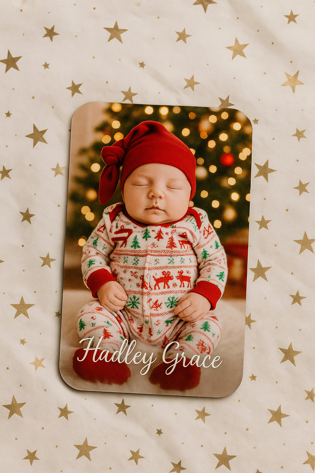 Baby in Christmas-themed outfit with red hat and patterned pajamas on a decorative card with gold stars and blurred lights.