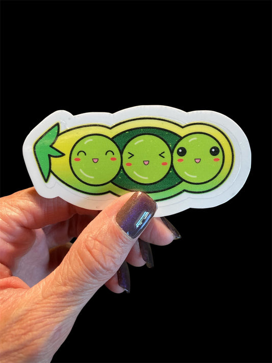 Hand holding a sticker with three cartoon peas with faces on a black background