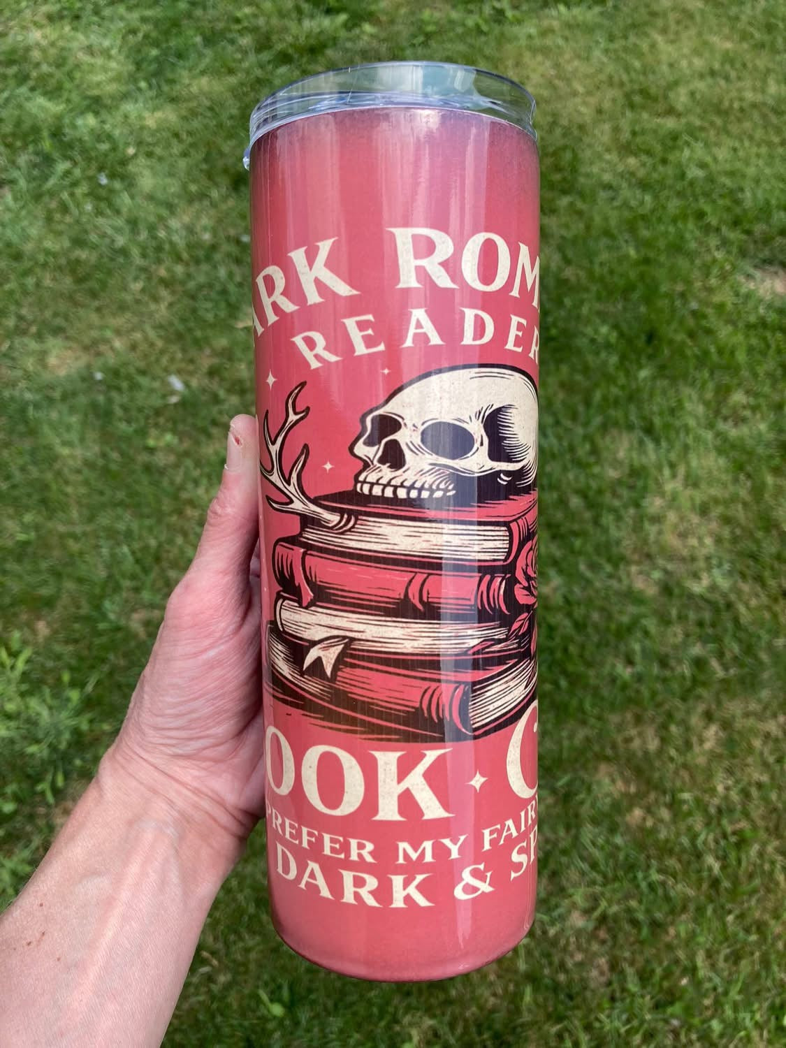 Pink tumbler with skull and books design held in front of grass. 20oz skinny tumbler with dark romance reader design featuring skull and stacked books on a red background