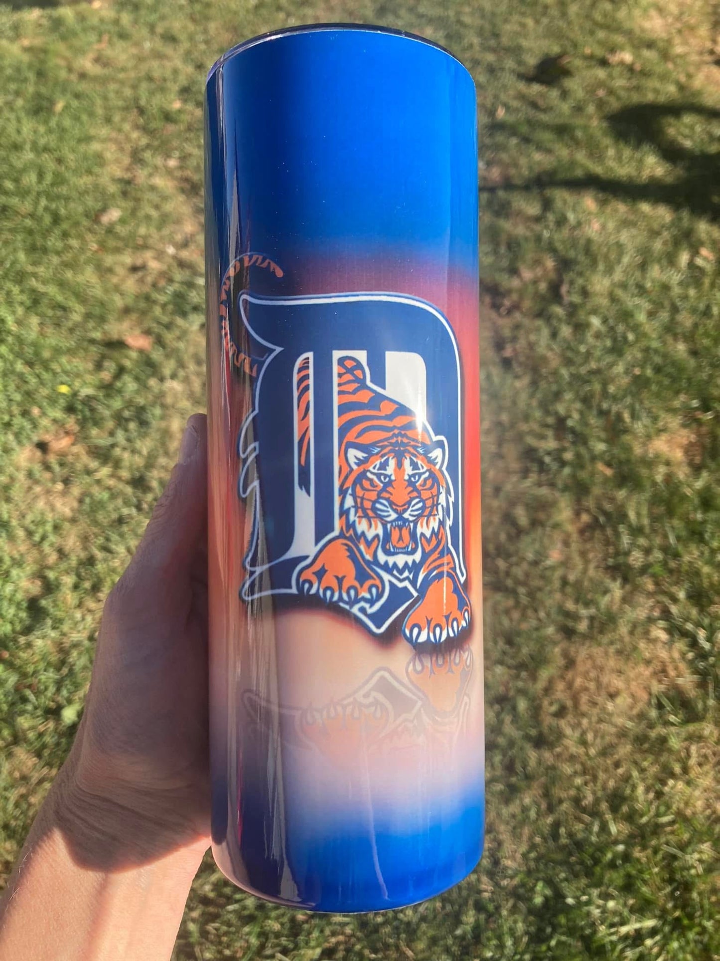 Tumbler with a tiger logo and blue gradient held in a hand on grass
