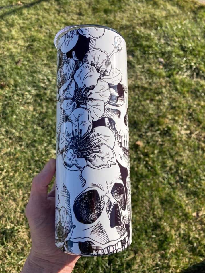 Tumbler with black and white floral and skull design held in front of grass. 20oz skinny tumbler featuring black and white skull with detailed floral design wrapping around the tumbler