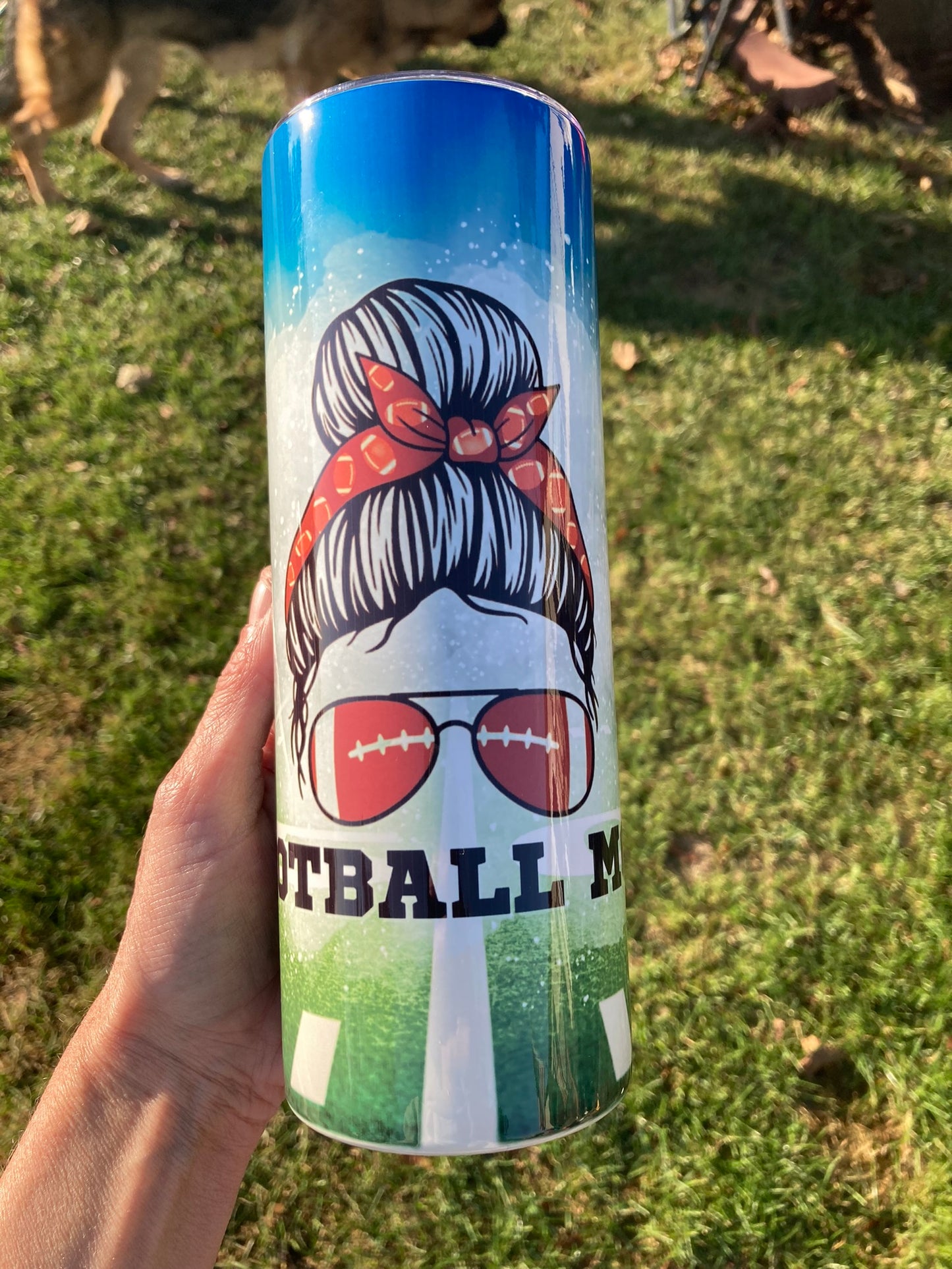 Tumbler with a design of a girl with sunglasses and 'Football Mom' text, held outdoors. 20oz skinny tumbler with football mom design featuring messy bun with red bandana and football sunglasses