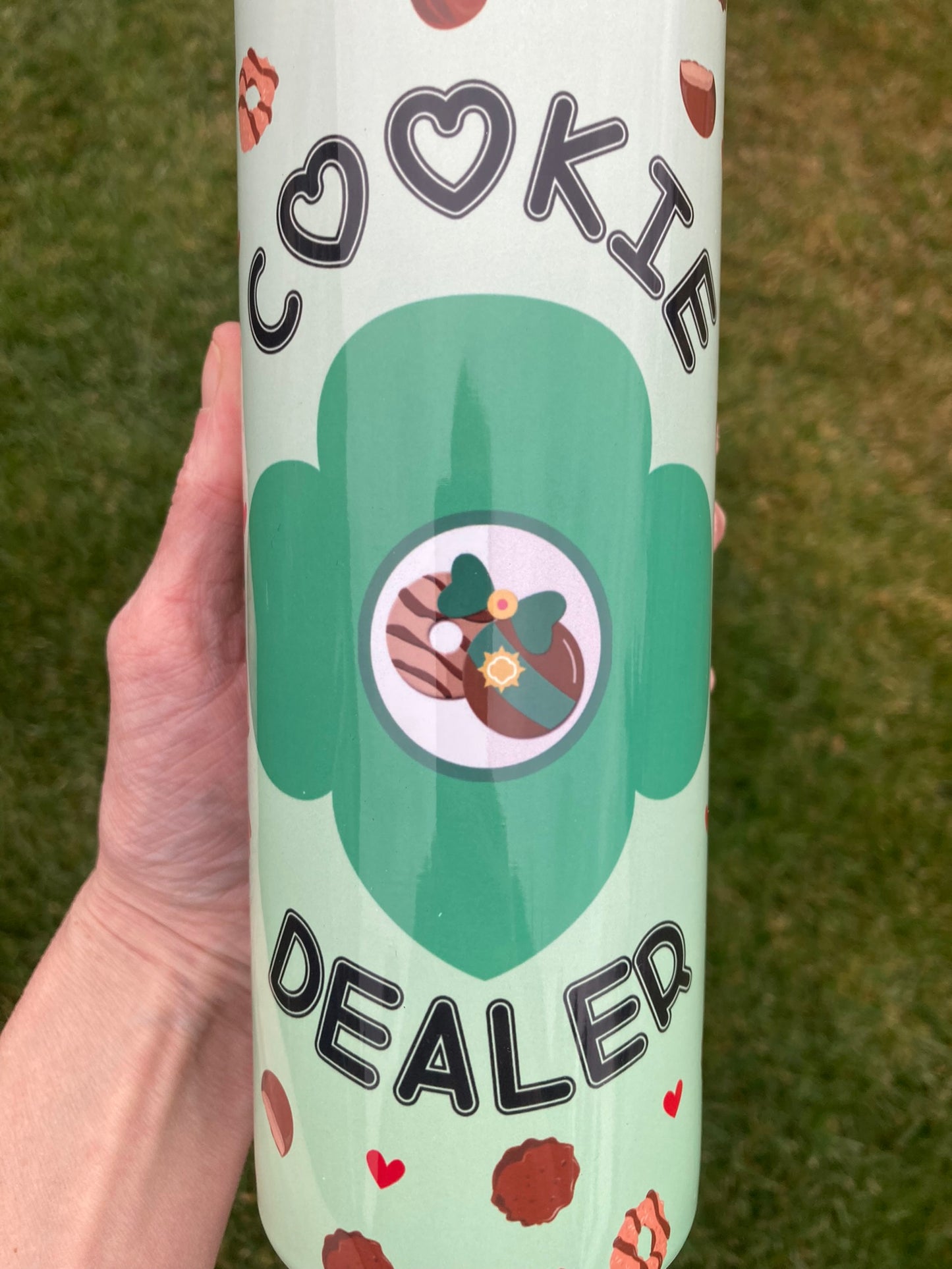 Hand holding a 20 oz Girl Scout Cookie Dealer stainless steel tumbler outdoors, showing green cookie-themed wrap with hearts and cookie illustrations during cookie season.