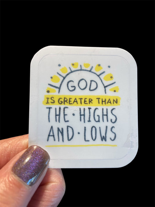 Hand holding a small card with 'God is greater than the highs and lows' text on a black background