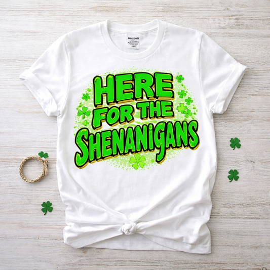 White t-shirt with green text 'HERE FOR THE SHENANIGANS' on a wooden surface with shamrock decorations. White unisex St. Patrick’s Day t-shirt with bold green “Here for the Shenanigans” text and shamrock graphics on Gildan 64000 tee.