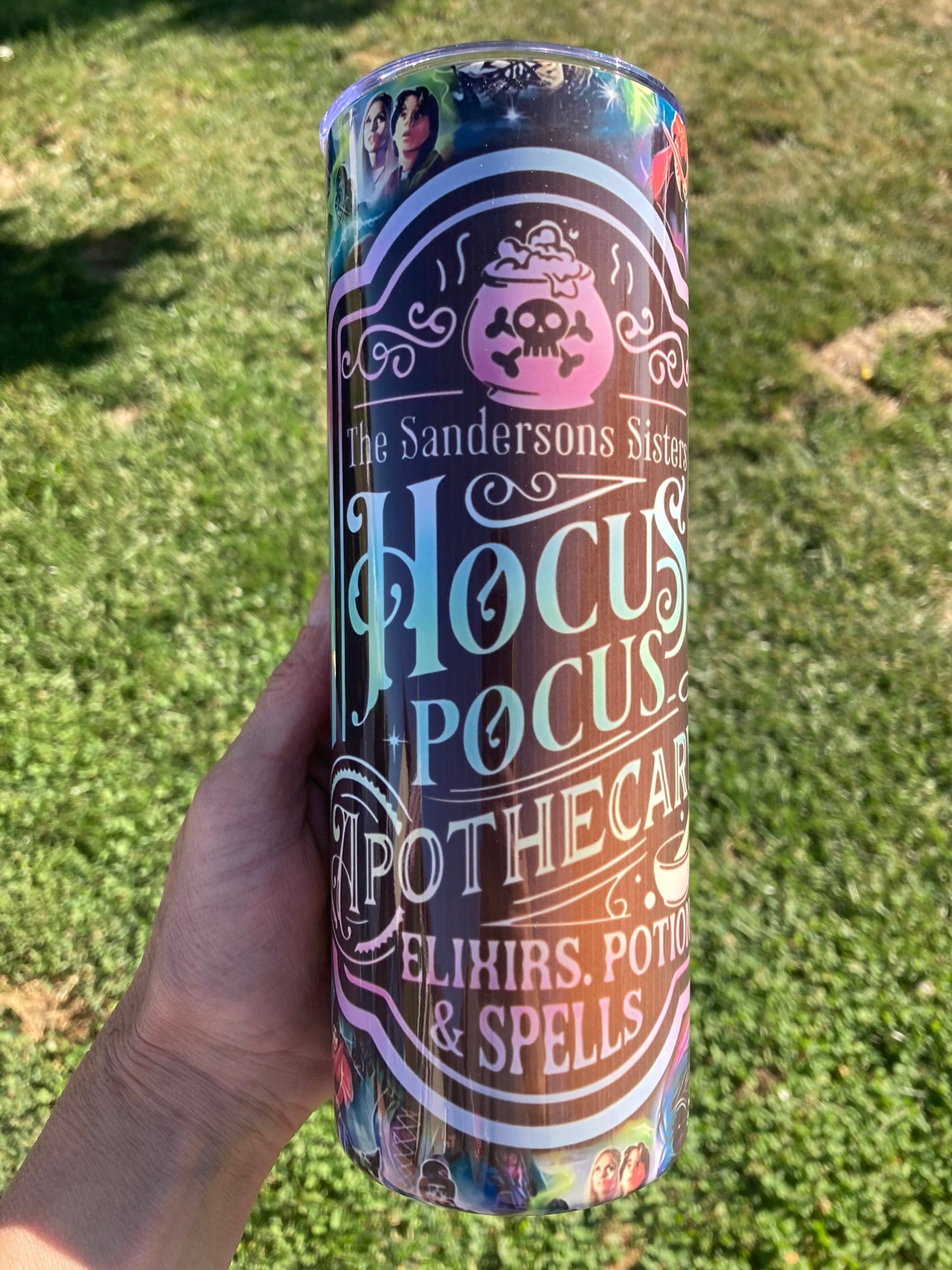 Tumbler with 'Hocus Pocus' design held in a hand against a grassy background. Hocus Pocus apothecary tumbler with potion bottle design and colorful witch themed background