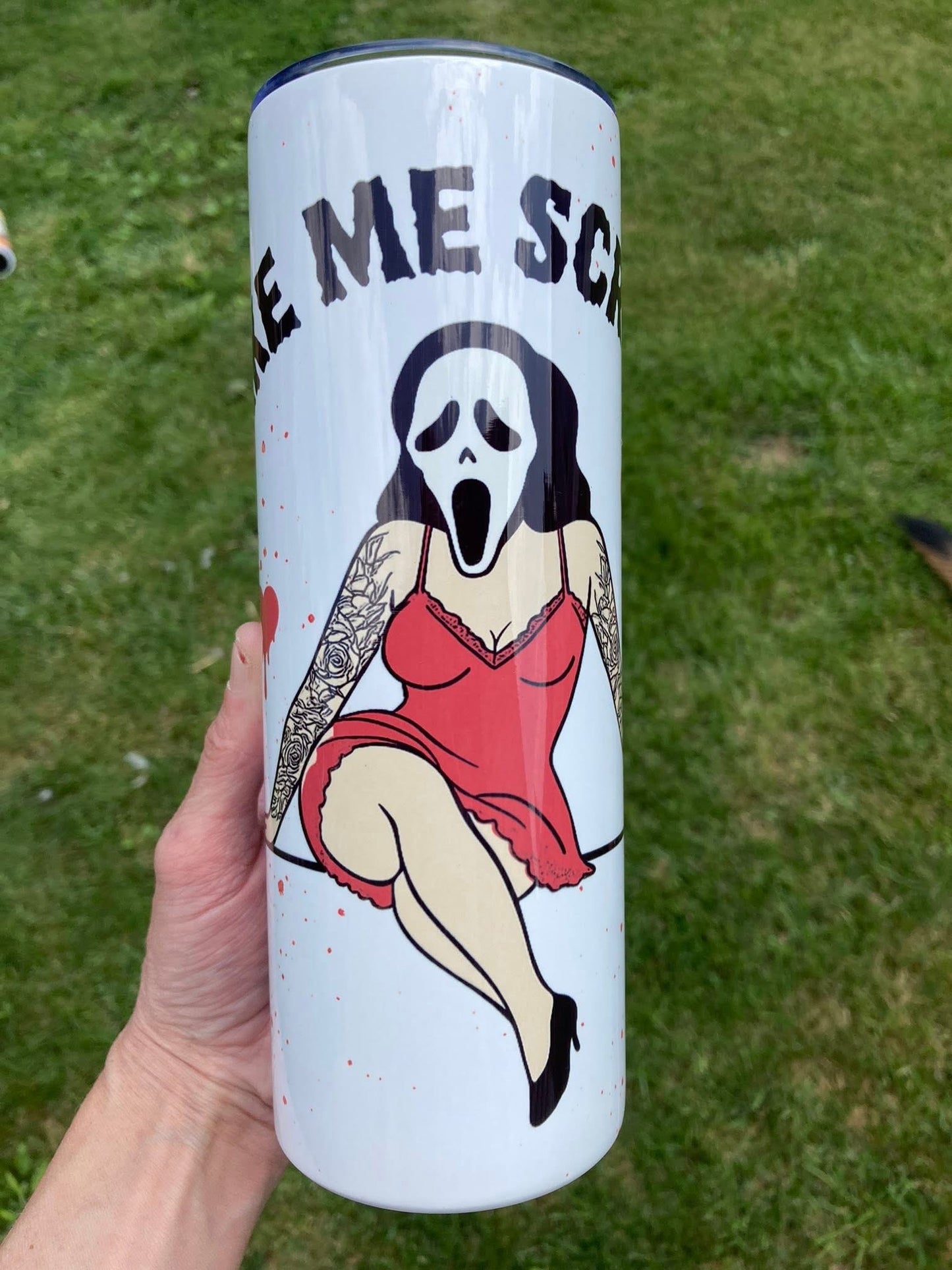 Tumbler with a graphic design of a woman in a red dress and mask on a grassy background. Horror scream girl tumbler featuring spooky masked character in red outfit with tattoo sleeves on white background