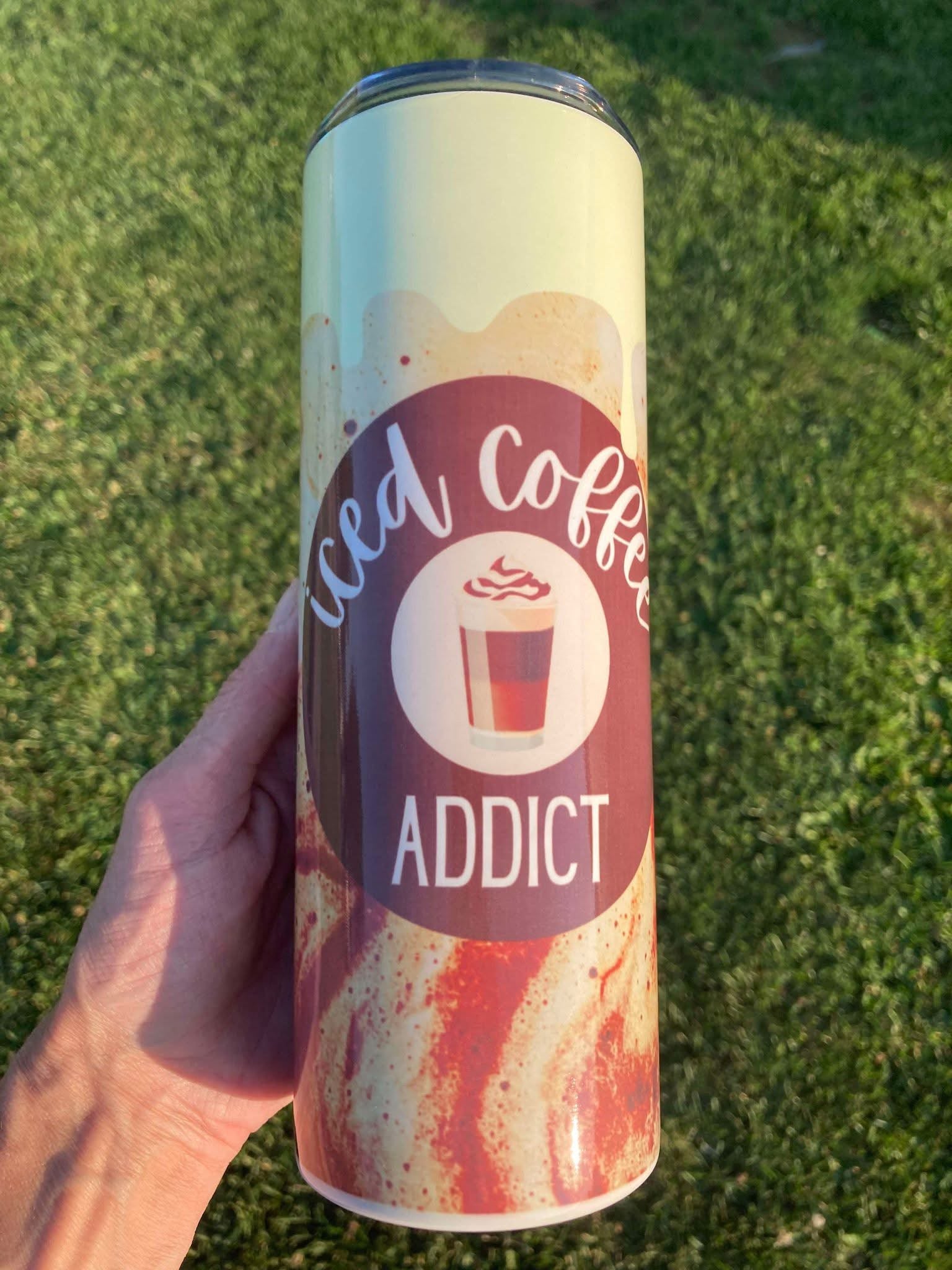 Tumbler with 'Iced Coffee Addict' design held against a grass background