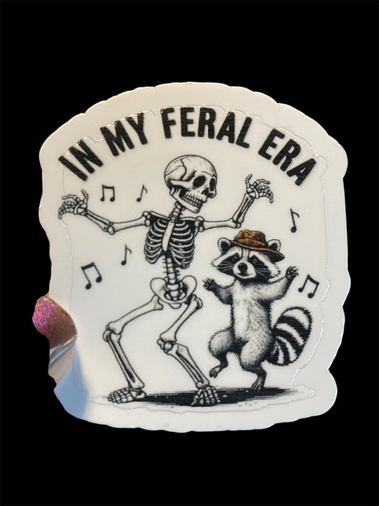 Sticker with skeleton and raccoon dancing, text 'In My Feral Era' on a black background