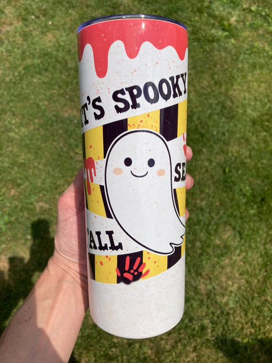 Tumbler with Halloween design held in front of grass. It’s Spooky Season tumbler with smiling ghost, yellow and black stripes, and red drip design for Halloween