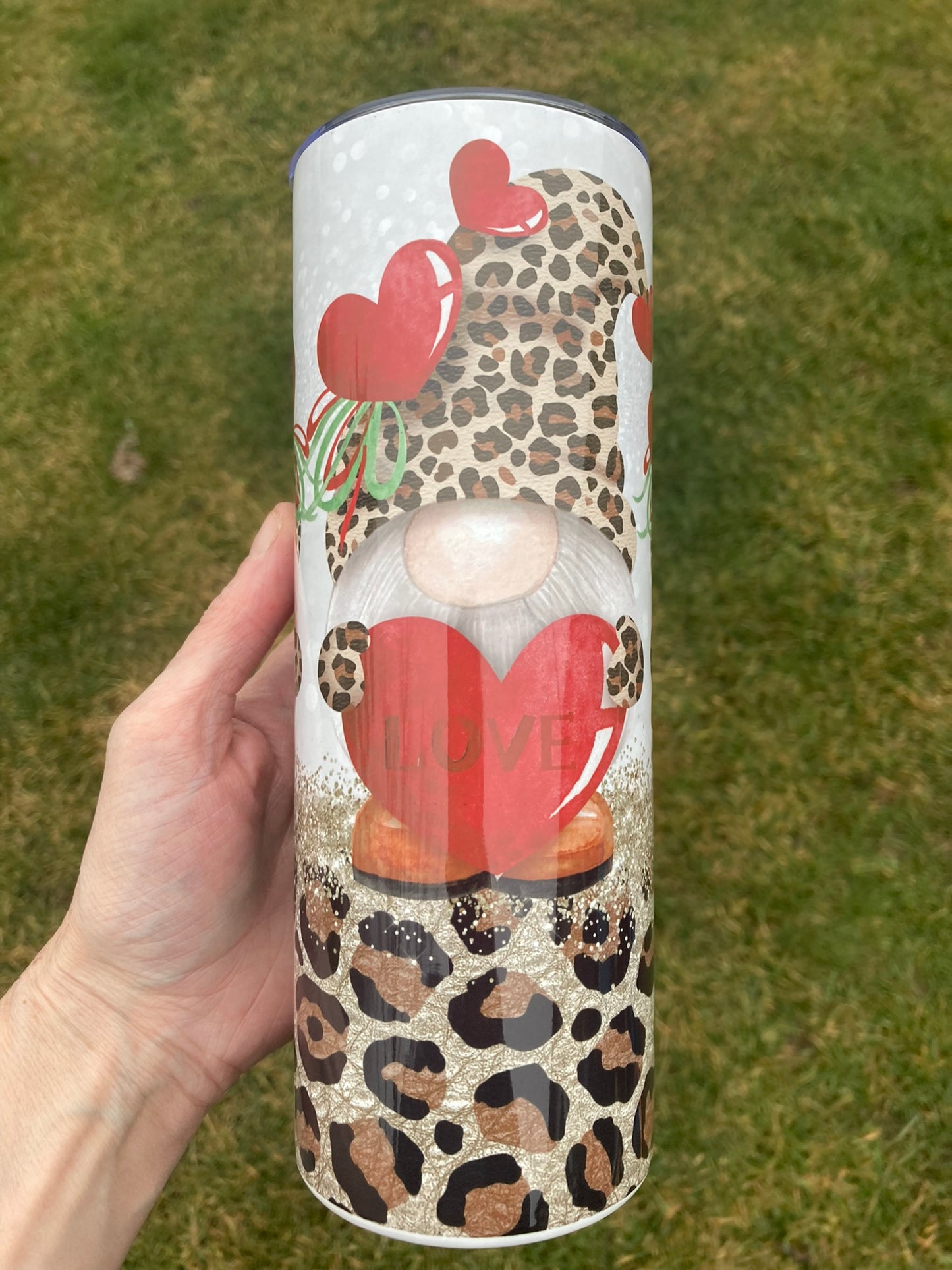 Tumbler with a design of a gnome holding a heart and leopard print pattern, held by a hand against a grassy background.