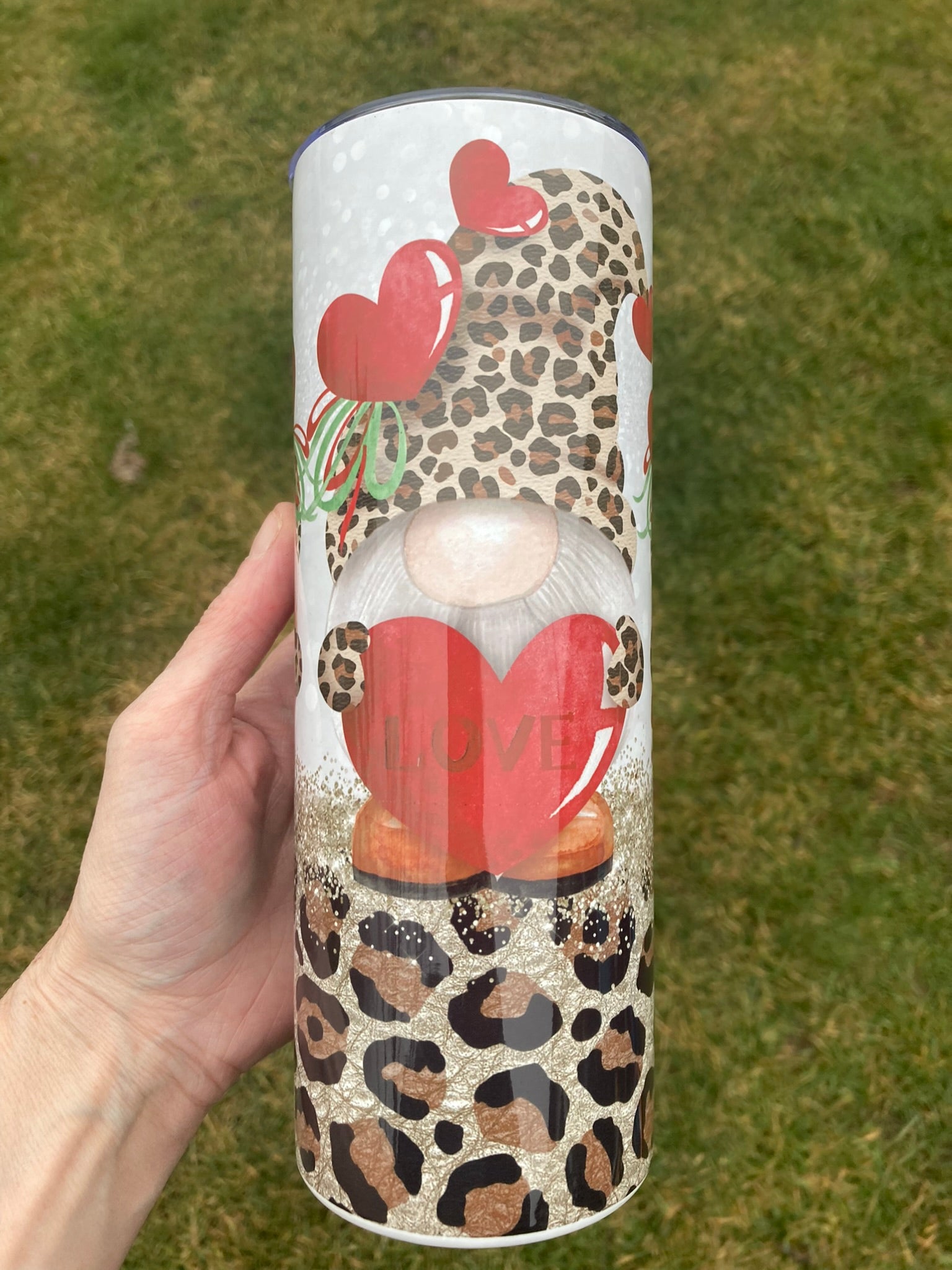 Tumbler with a design of a gnome holding a heart and leopard print pattern, held by a hand against a grassy background.