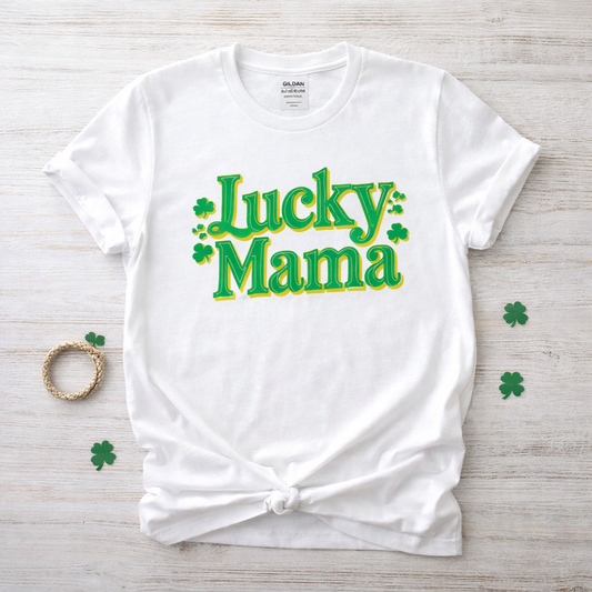 White t-shirt with 'Lucky Mama' text on a wooden surface with shamrock decorations. White Lucky Mama St. Patrick’s Day shirt with green and yellow lettering and shamrock accents.