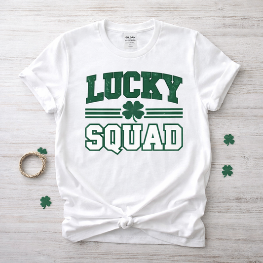 White t-shirt with 'Lucky Squad' text and shamrock design on a wooden surface. White Lucky Squad St. Patrick’s Day t-shirt with green varsity style lettering and shamrock graphic.