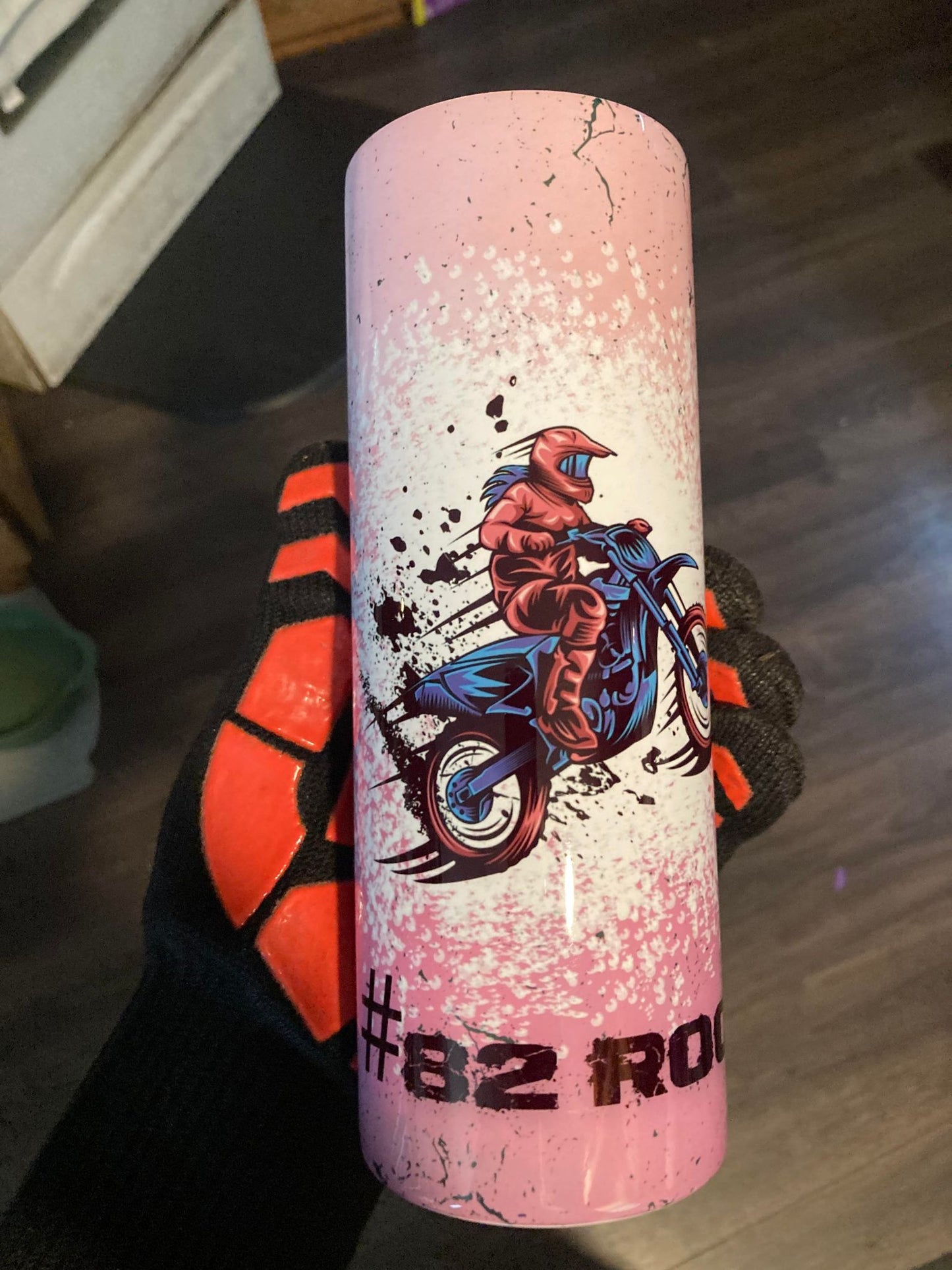 Pink water bottle with graphic design of a person on a motorcycle, held by a person wearing black and orange shoes.