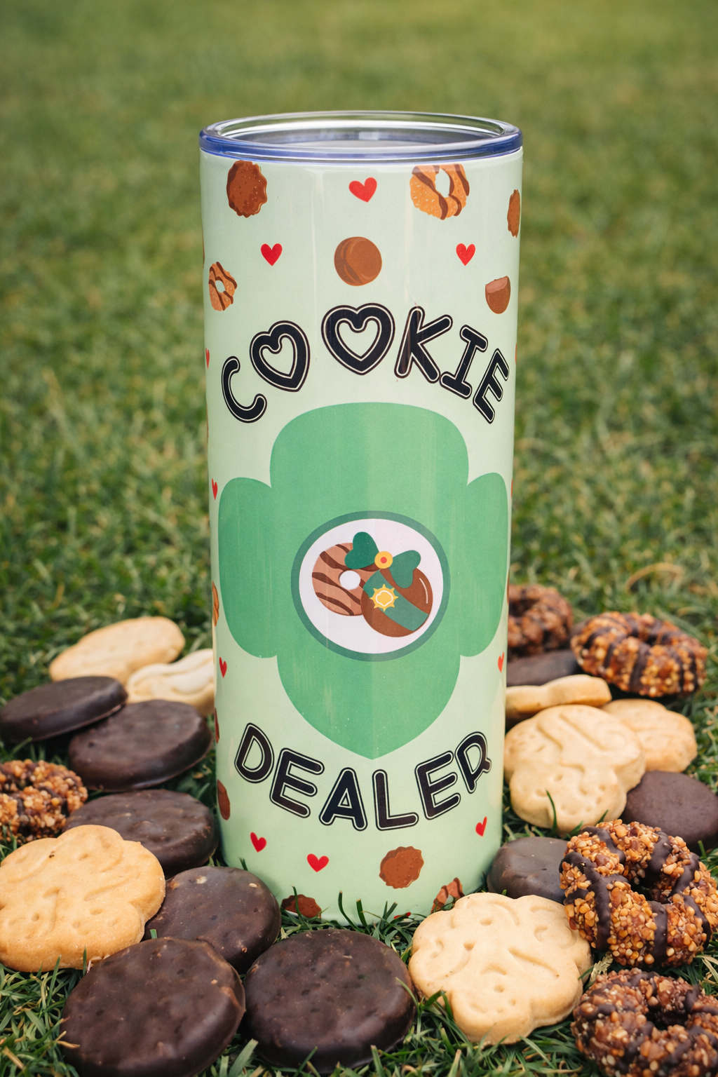 Girl Scout Cookie Dealer 20 oz stainless steel tumbler standing on grass, surrounded by assorted Girl Scout cookies including chocolate-covered and shortbread styles, featuring a green cookie-themed design with hearts.