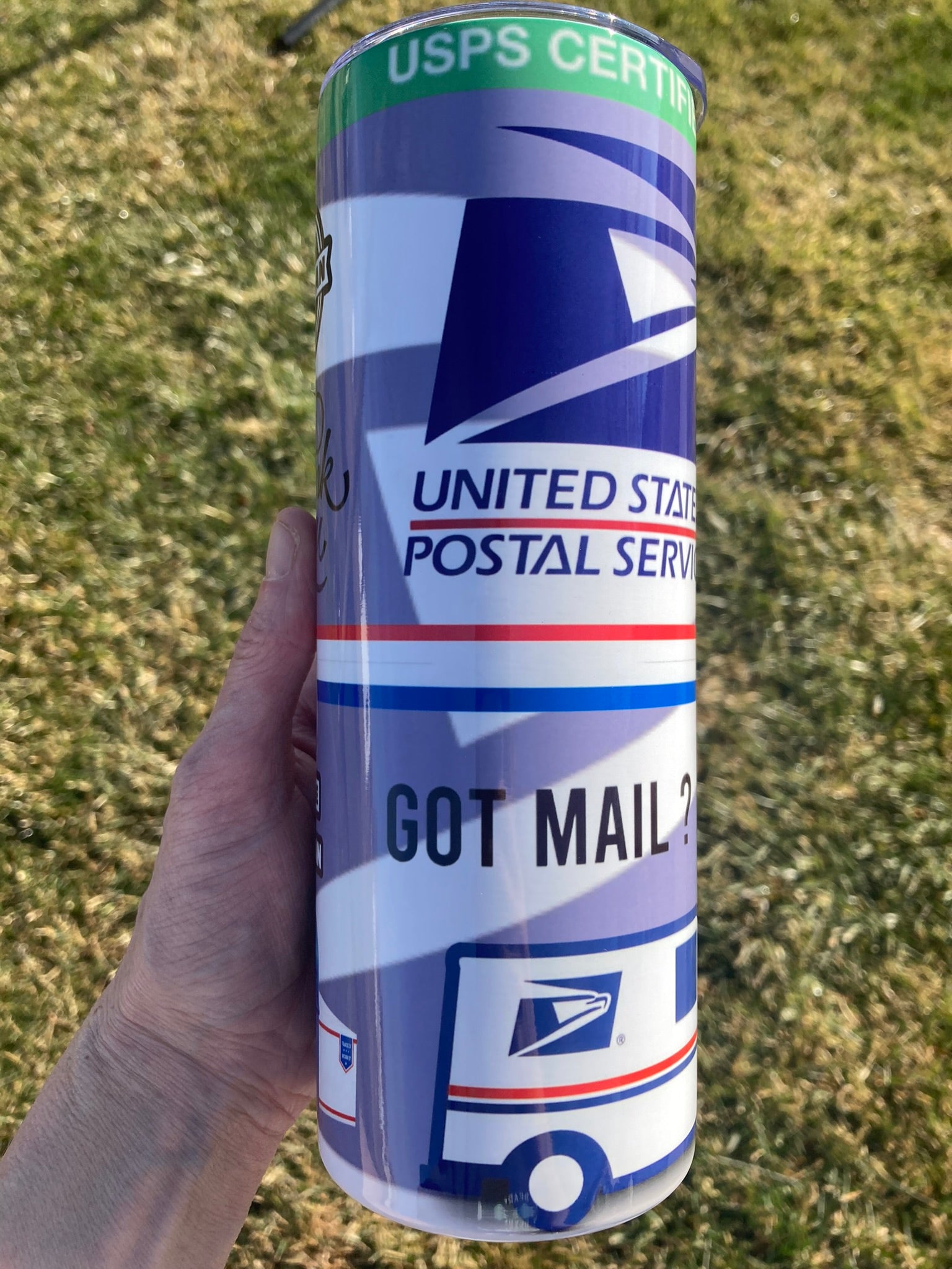 Hand holding a cylindrical object with United States Postal Service branding on a grassy background