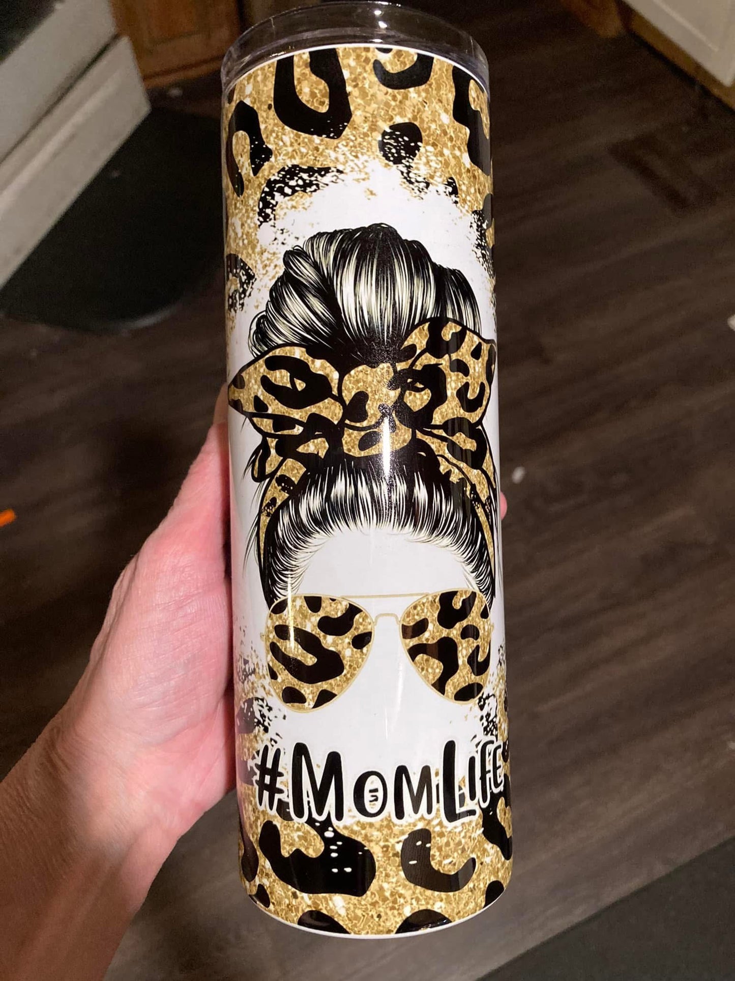 Tumbler with leopard print and 'Mom Life' text held by a hand on a wooden floor. 20oz skinny tumbler with messy bun mom design featuring leopard print bow and sunglasses with hashtag momlife text