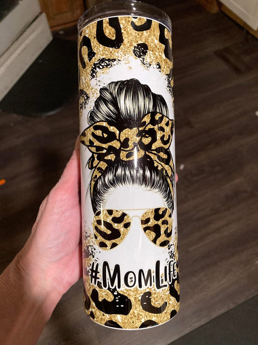 Tumbler with leopard print and 'Mom Life' text held by a hand on a wooden floor. 20oz skinny tumbler with messy bun mom design featuring leopard print bow and sunglasses with hashtag momlife text