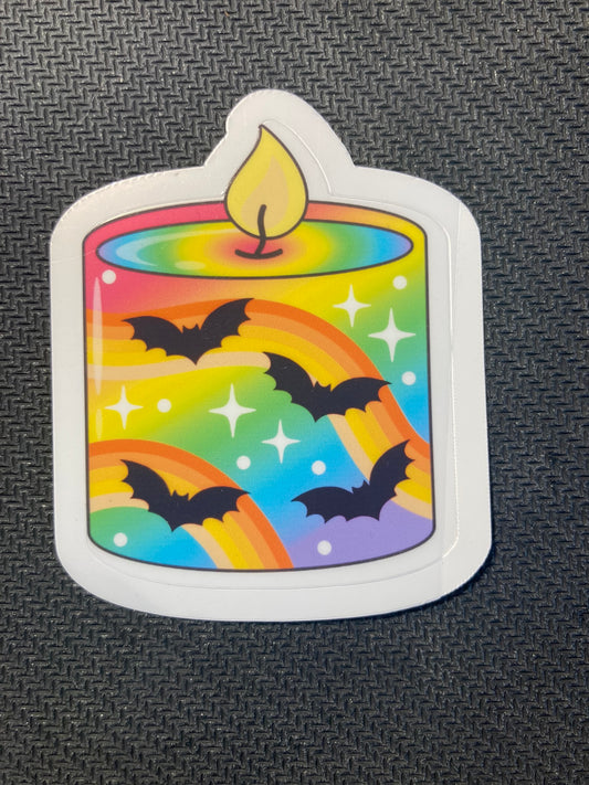 Halloween sticker of a rainbow-striped lit candle with black bats flying around sparkles.
