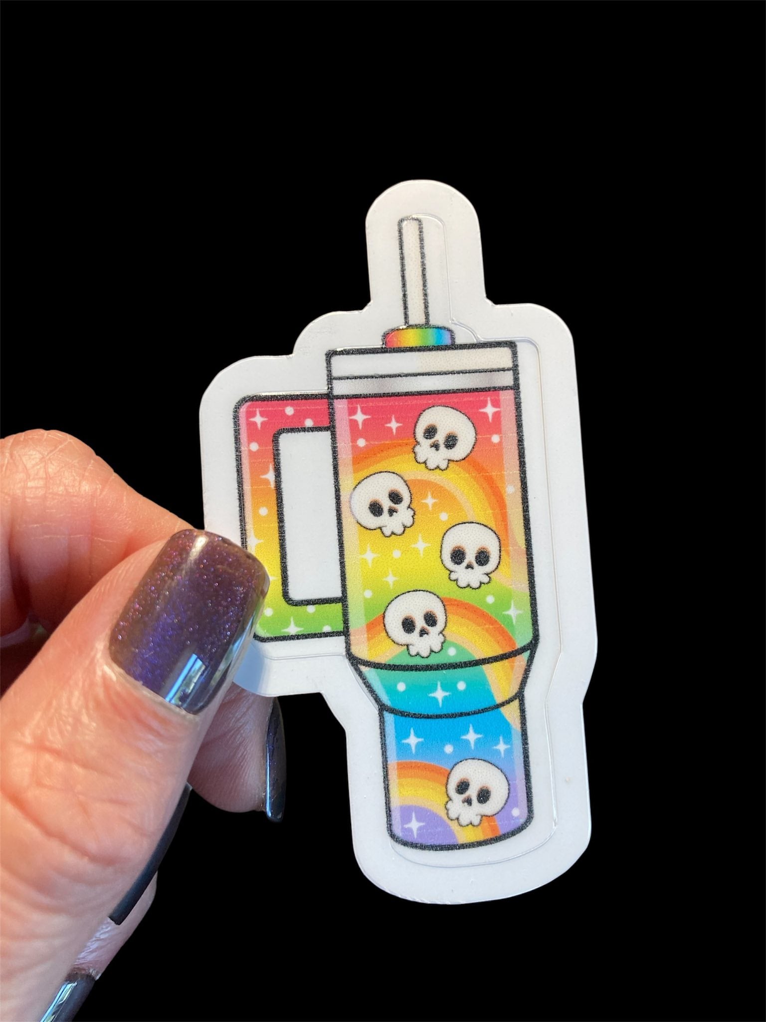 Sticker of a rainbow-themed mug with skull designs held by a hand against a black background