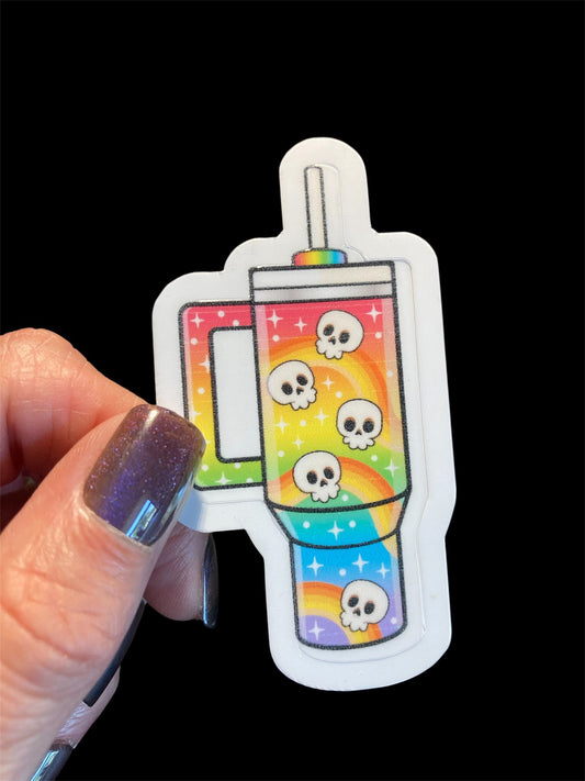 Sticker of a rainbow-themed mug with skull designs held by a hand against a black background