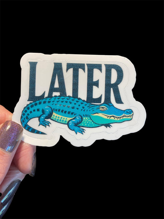 Sticker with an alligator and 'LATER' text held by a hand on a black background
