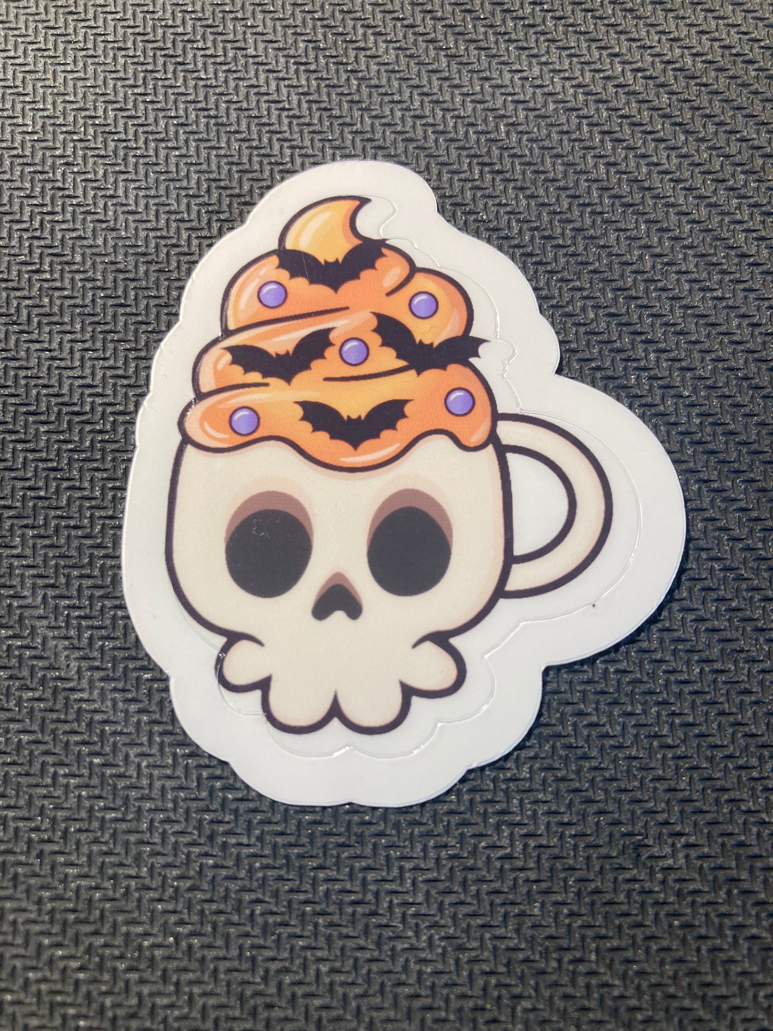 Halloween sticker of a skull-shaped coffee mug filled with orange whipped topping, bats, and sprinkles.